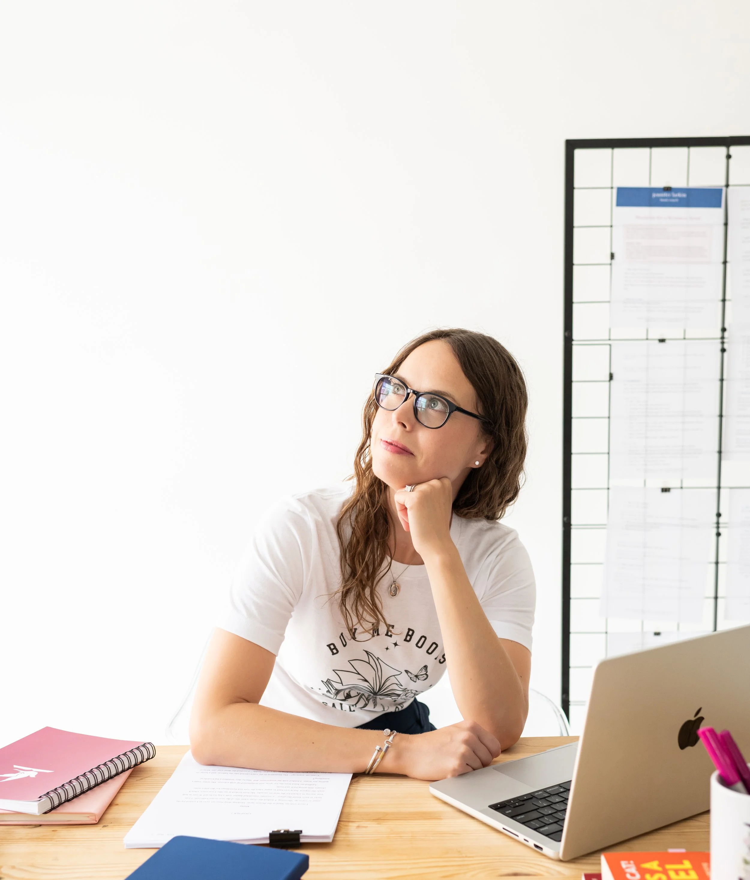 A woman with glasses sitting at a desk, resting her chin on her hand, looking thoughtfully upwards. On her desk are a laptop, notebooks, and papers. There is a black wire grid with papers posted on it in the background.