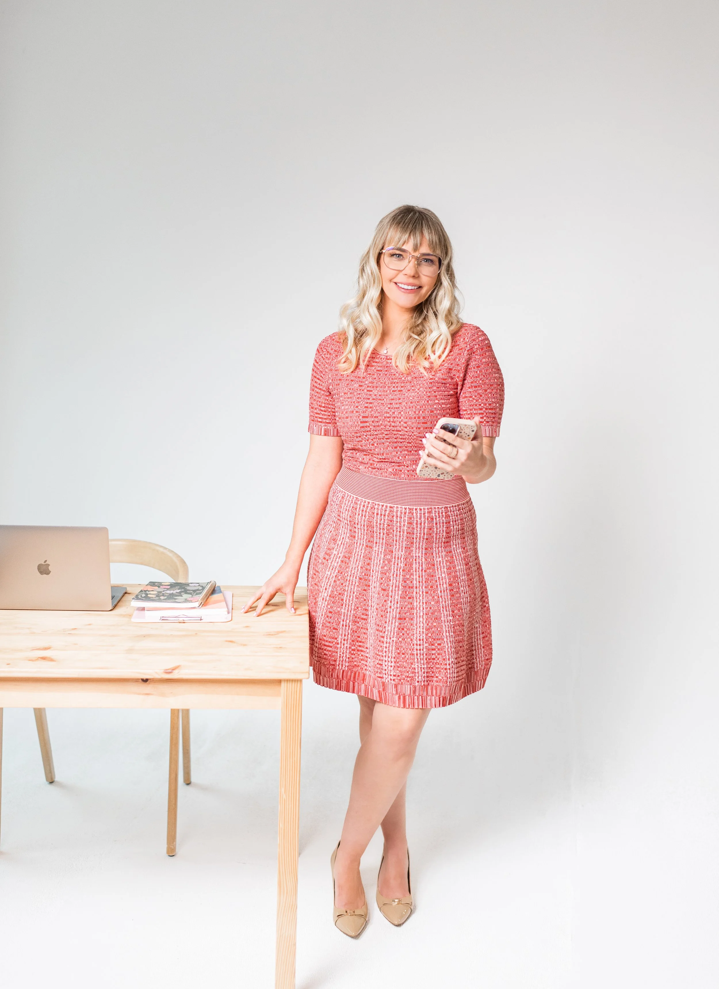A woman in a pink dress standing next to a wooden table holding a smartphone, with laptop and notebooks on the table, against a plain white background.