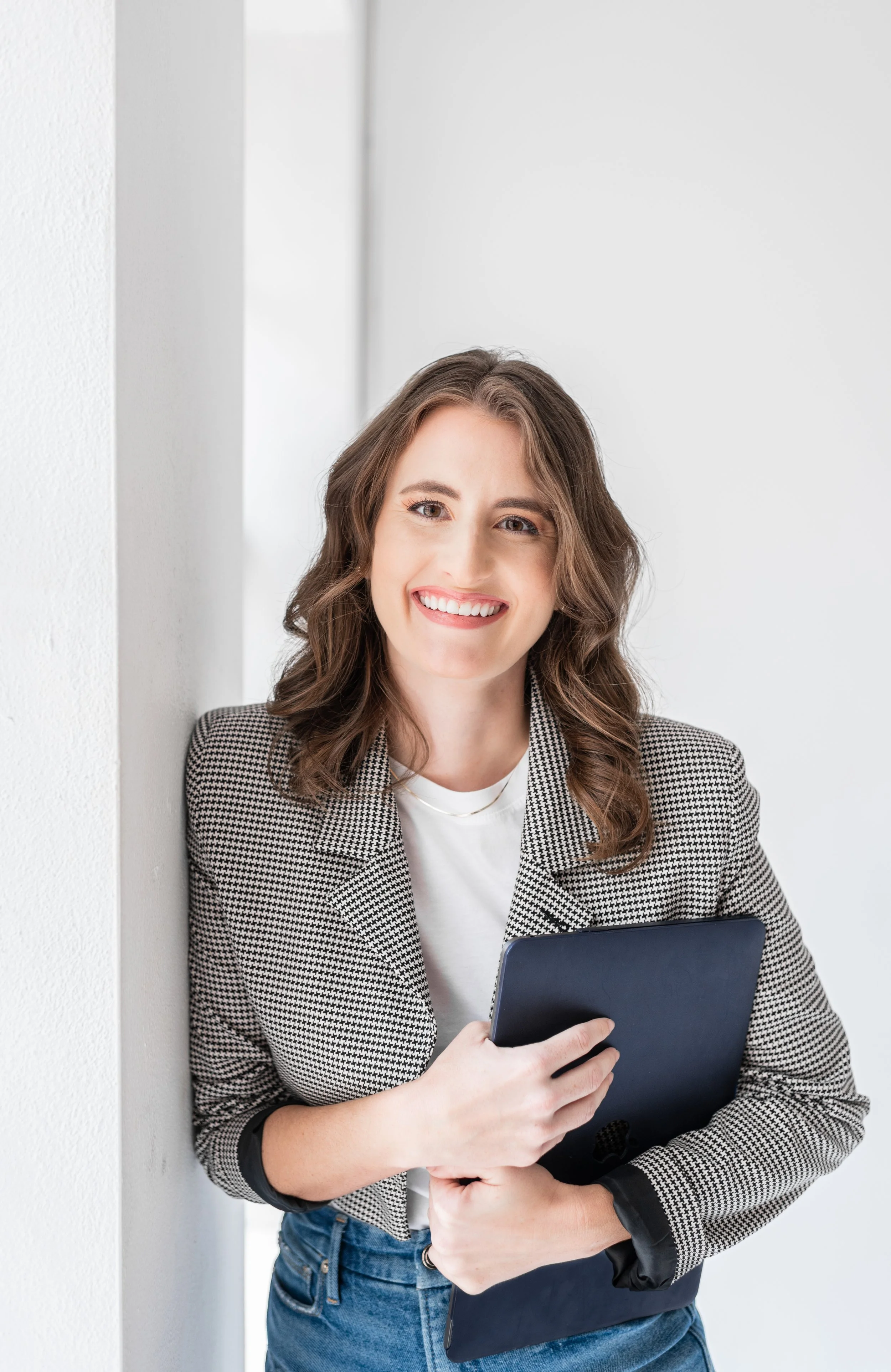 Woman smiling while holding a tablet, wearing a checkered blazer and casual jeans, standing against a white wall background.