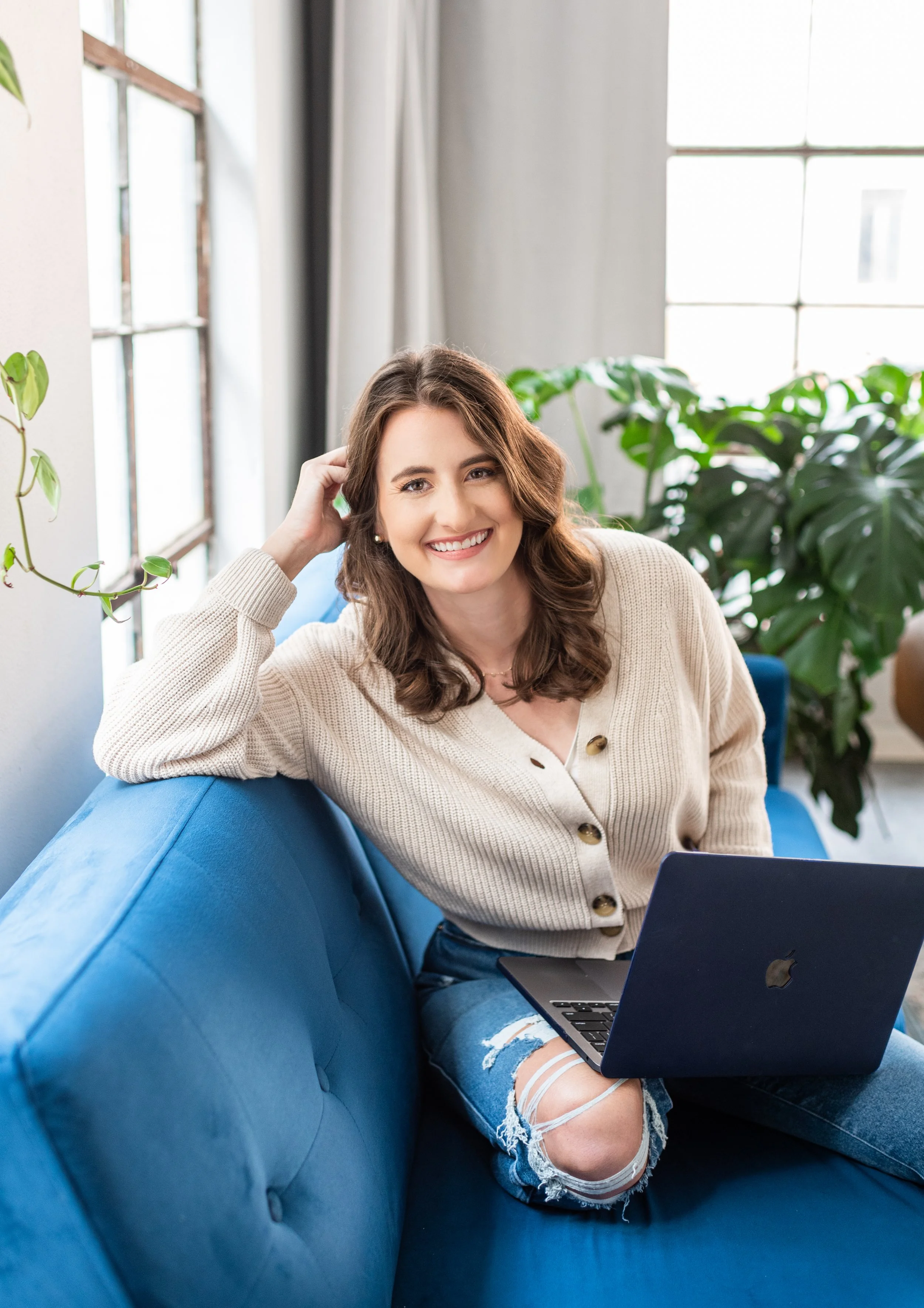 A woman with brown hair smiling while sitting on a blue sofa with a laptop on her lap in a bright, plant-filled room.