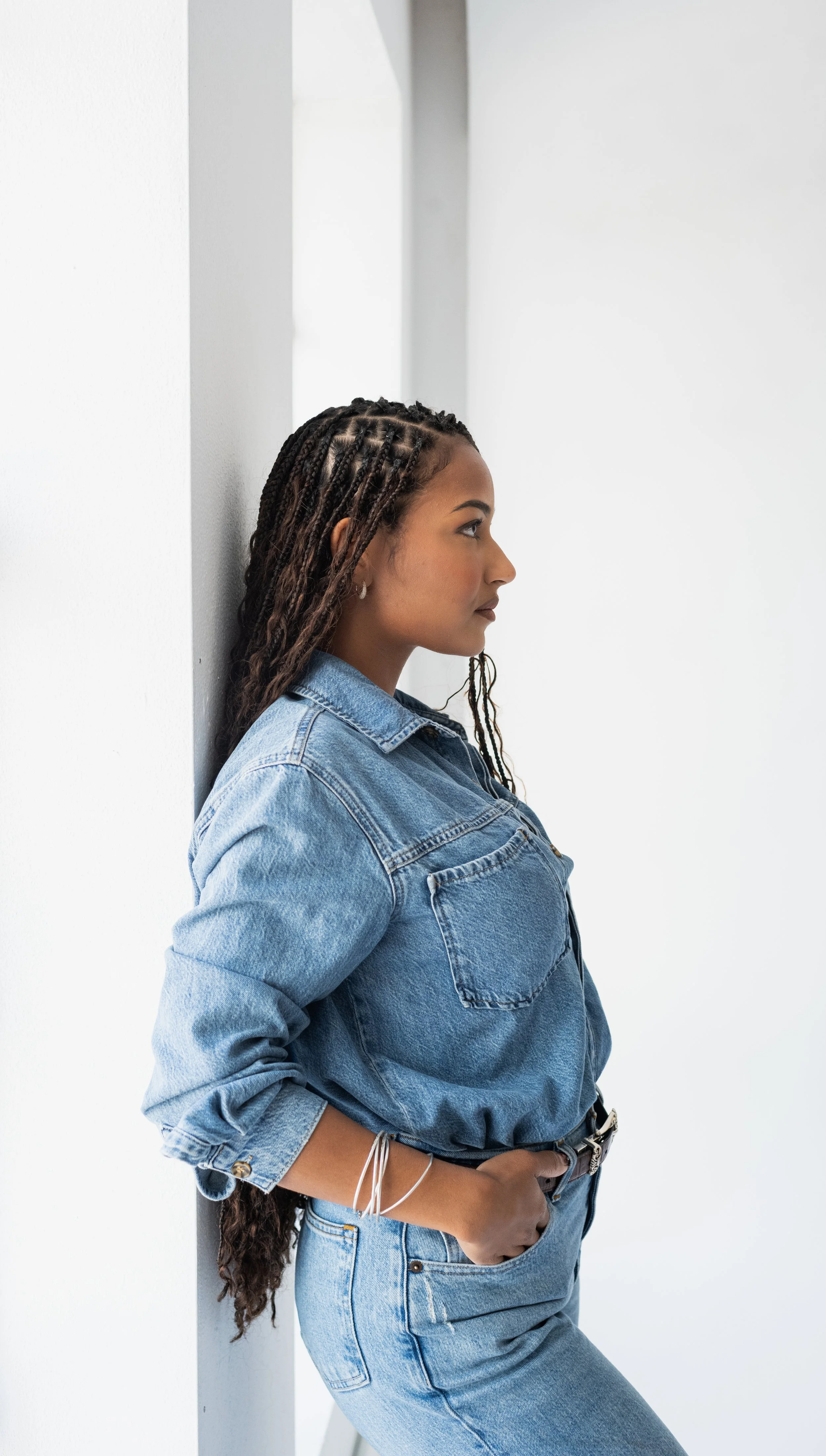 A woman with braided hair and hoop earrings, wearing a denim jacket and jeans, leaning against a white wall with her hands in her pockets.