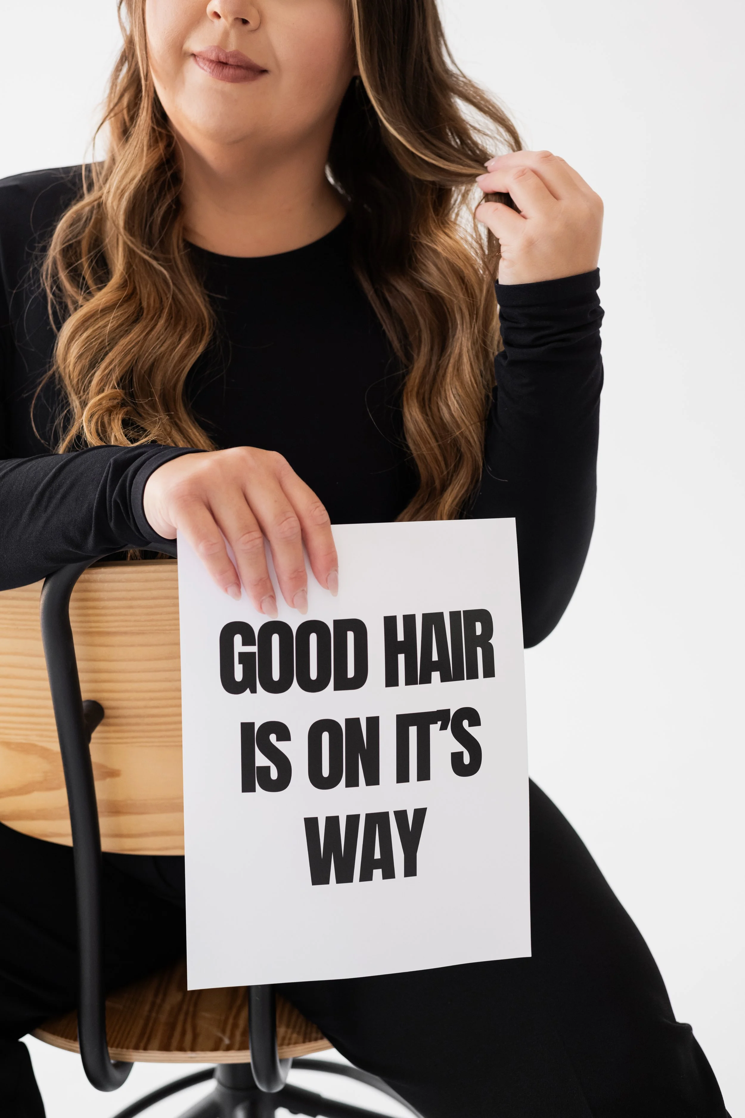 Woman sitting on a wooden chair, holding a sign that says 'Good hair is on its way' with black bold text, dressed in black, with long wavy brown hair, against a plain white background.