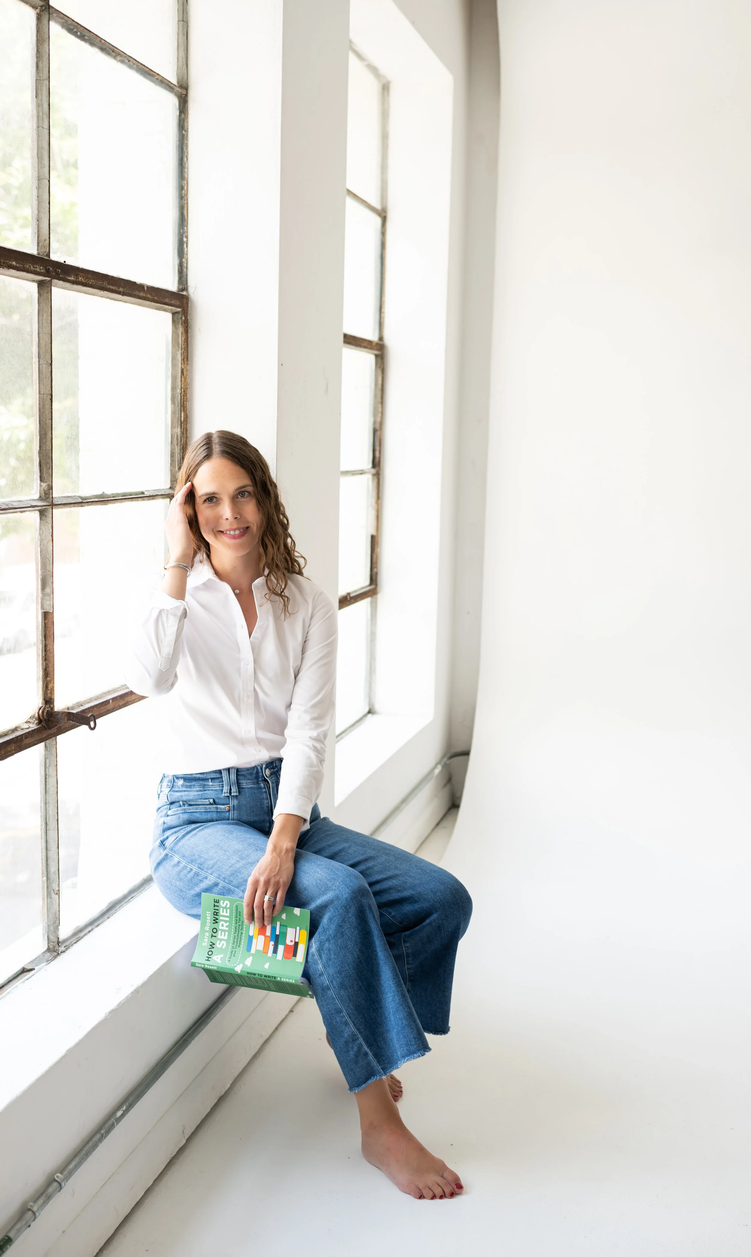 A woman with wavy brown hair wearing a white button-up shirt and blue jeans sitting on a wide window sill, holding a book titled "How to Write a Series" in her lap, smiling and touching her hair.