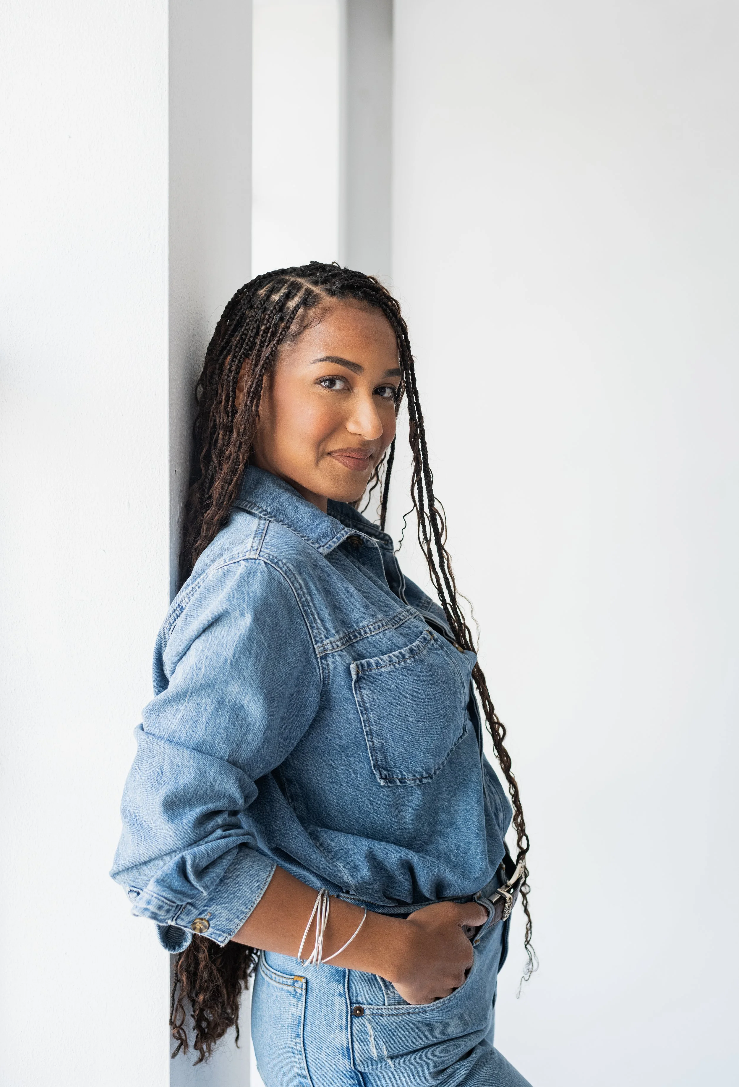 A young woman with long dreadlocks leaning against a white wall, wearing a denim jacket and jeans, looking at the camera with a slight smile.