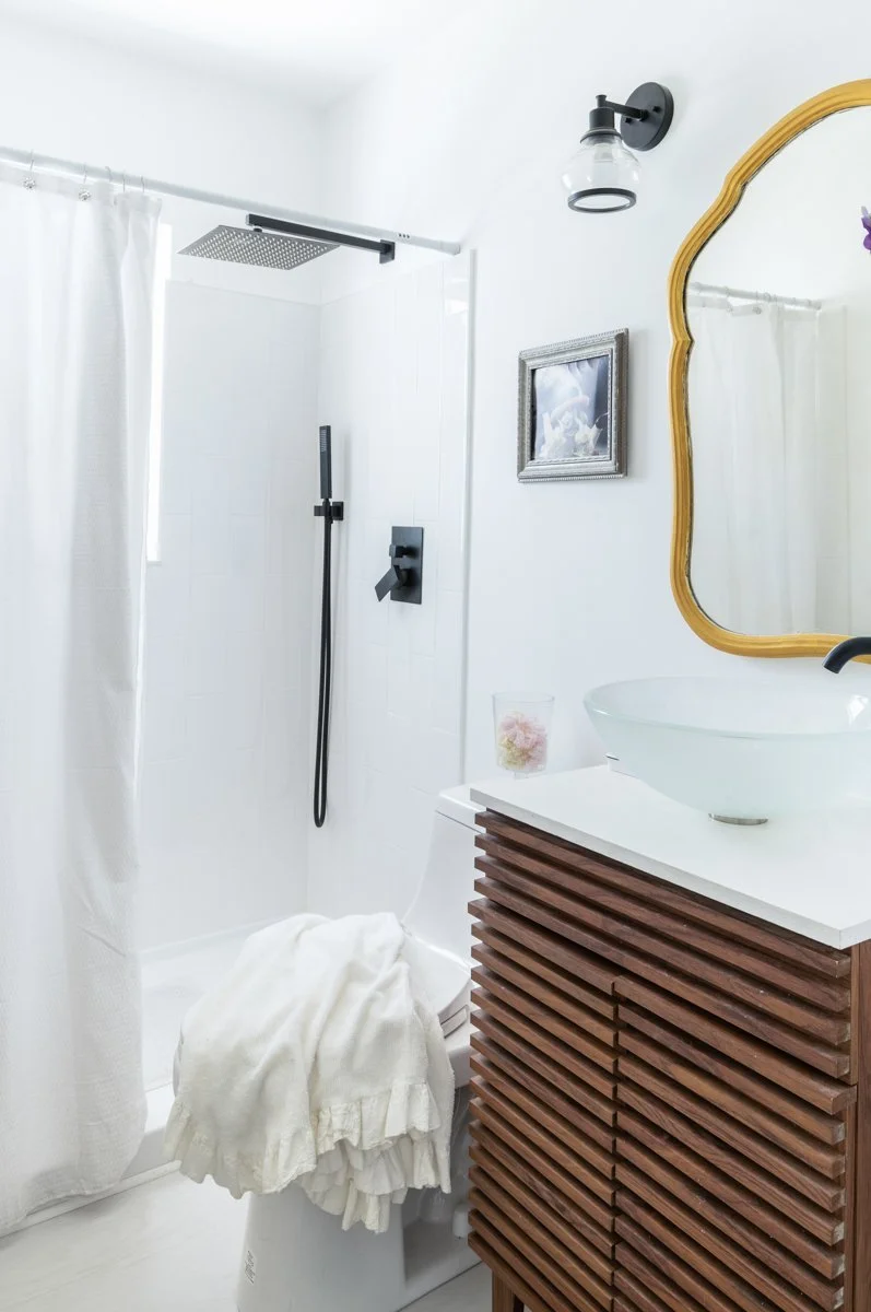 Modern bathroom with walk-in shower, white walls, black fixtures, wooden vanity with vessel sink, gold-framed mirror, small framed picture, and soft lighting.