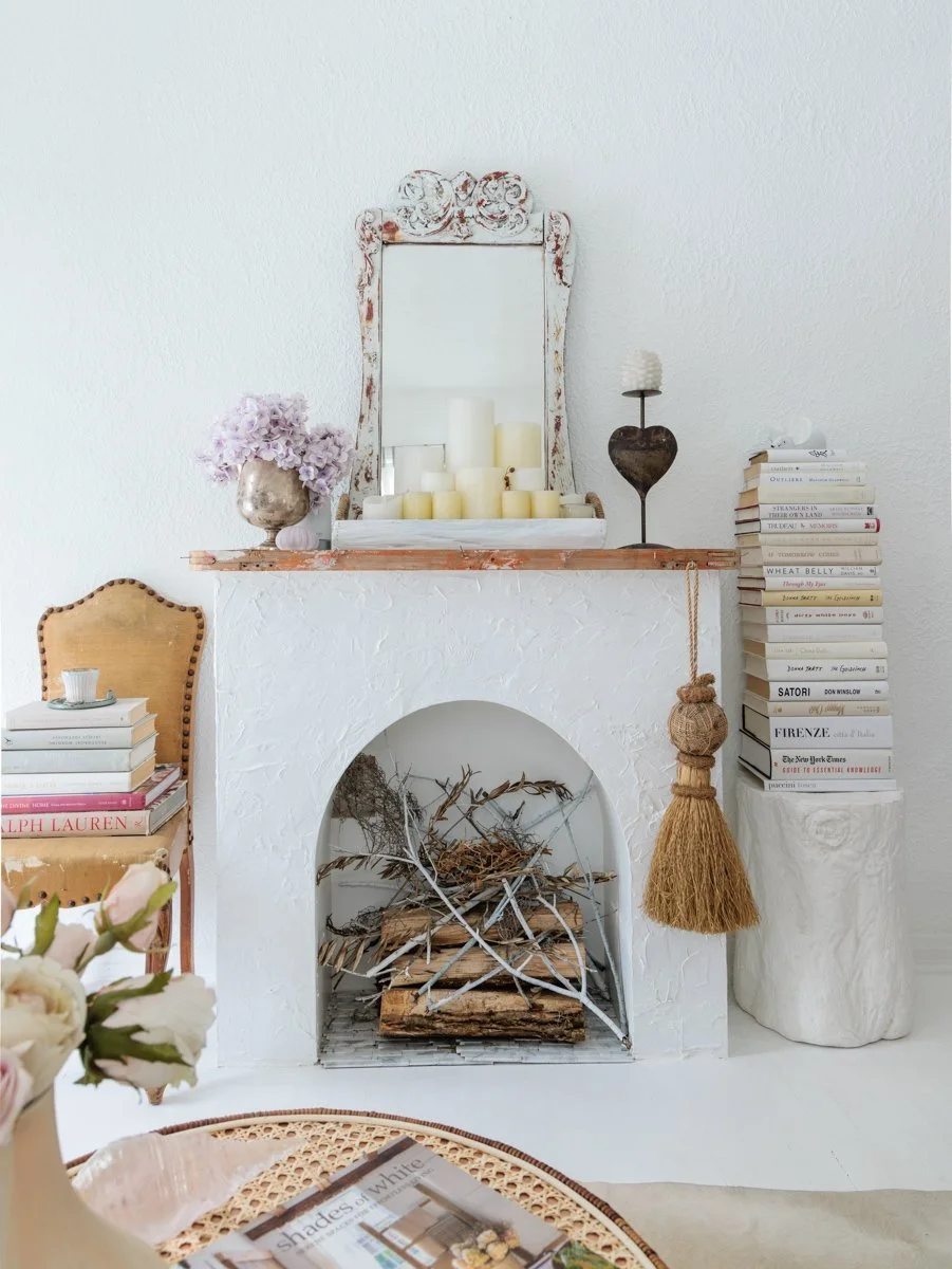Decorative white fireplace with a mirror above, surrounded by flowers, candles, and stack of books; including a chair with books and a teacup, and a vase of flowers in the foreground.