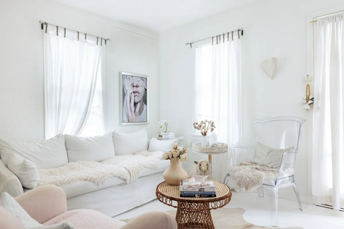 Bright white living room with white sofa, framed artwork, sheer curtains, a clear chair with a cover, and decorative accessories in neutral tones.