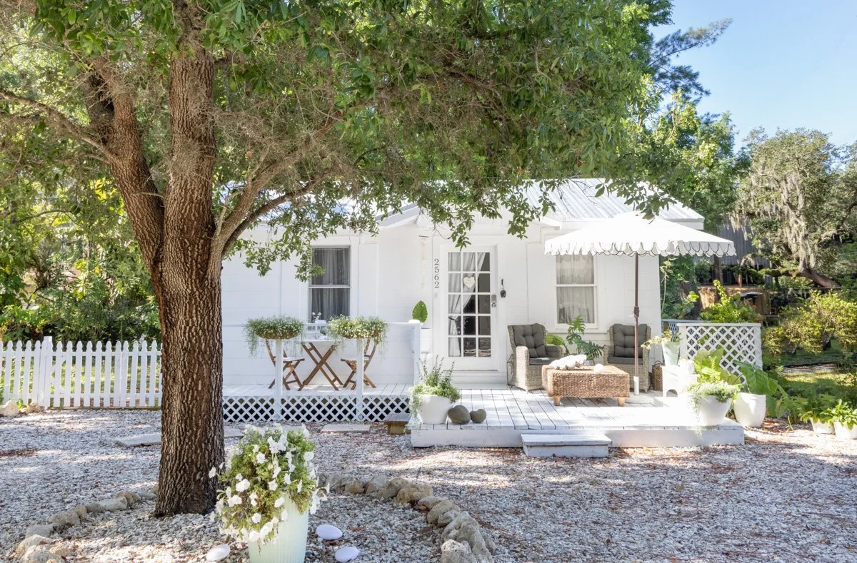 White house with porch, surrounded by trees and plants, outdoor seating with cushions and a white umbrella, gravel yard with flower pots.
