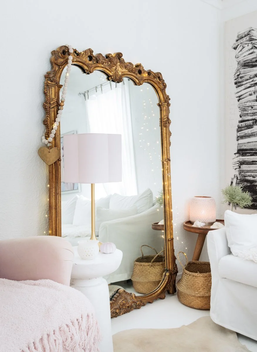 A cozy living room with a tall, ornate gold-framed mirror leaning against the wall. The mirror reflects a white couch and curtains. Decor includes a white table with a table lamp, a woven basket, and string lights around the mirror. There's artwork o