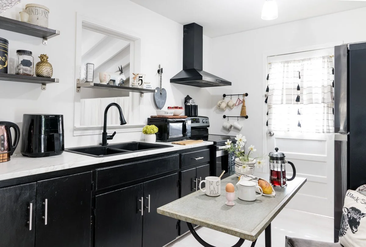 A modern kitchen with black cabinets, white countertops, and stainless steel appliances. There's a small table with breakfast items, a vase with white flowers, and a window with striped curtains.