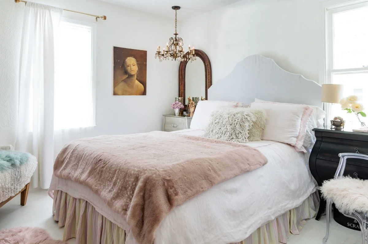 A cozy bedroom with white walls and natural light from two windows. The bed has a white headboard, pink pillows, and a fluffy cream pillow, covered with a white duvet and a light pink blanket. There are nightstands with a lamp, flowers, and decorativ