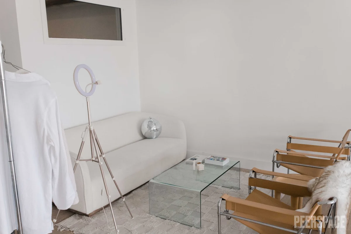 Modern living room with white sofa, glass coffee table, and two wooden chairs. A ring light on a tripod and a silver decorative object on the sofa. A clothing rack with white garment on the left and a fluffy white throw on one chair. Minimalist decor