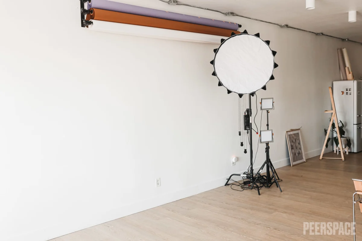 Photography studio with lighting equipment, including a large circular Softbox, two smaller LED lights on tripods, a backdrop roller system near the wall, a framed picture leaning against the wall, and a refrigerator in the corner.