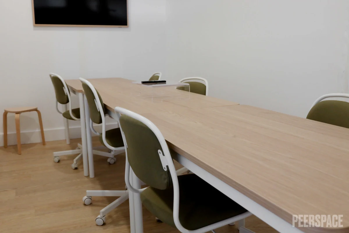 Empty conference room with a large wooden table, six green office chairs with white frames, a small wooden stool, and a wall-mounted black screen.