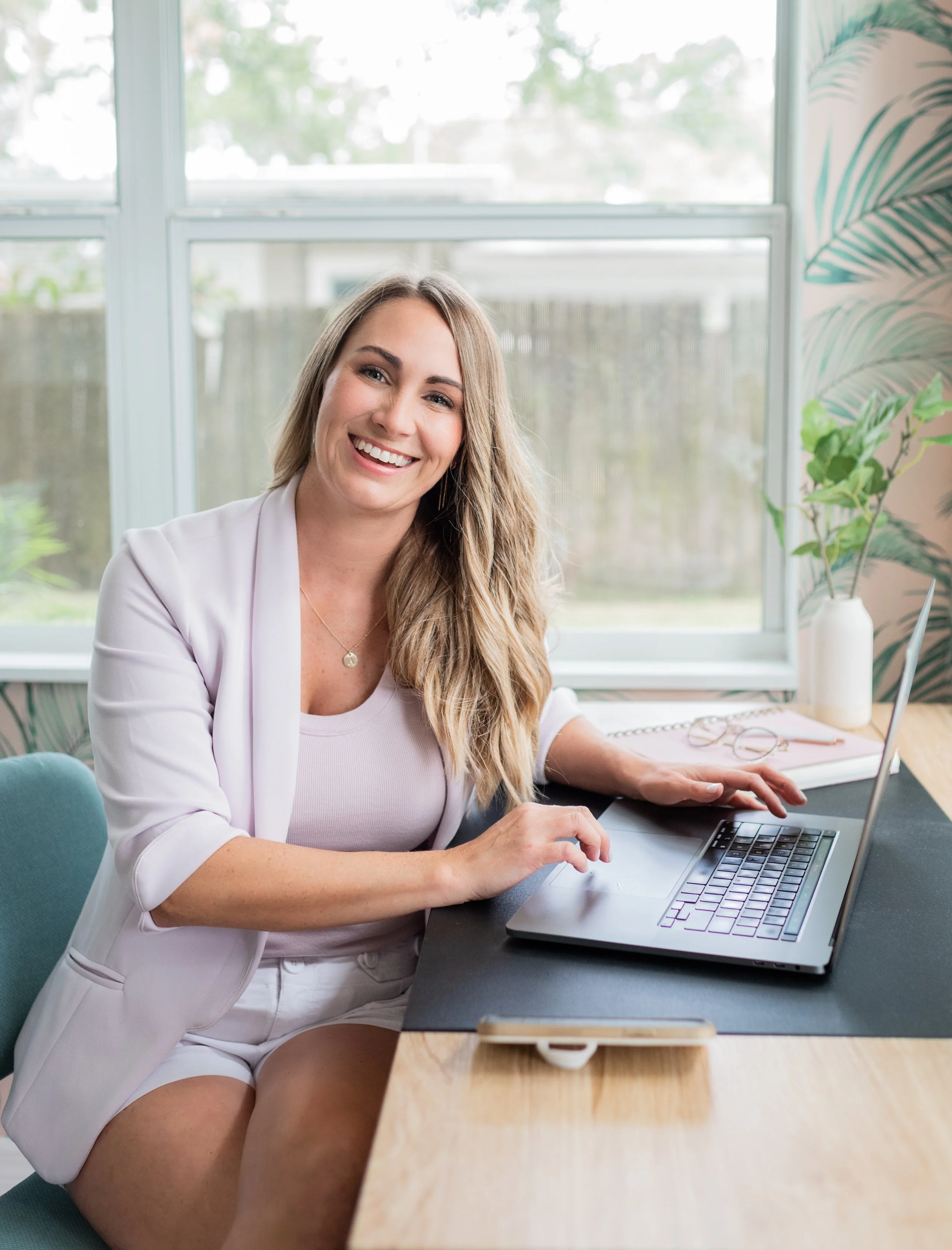 A woman with long blonde hair, smiling, sitting at a desk in front of a laptop, in a room with large windows and leafy plants in the background.