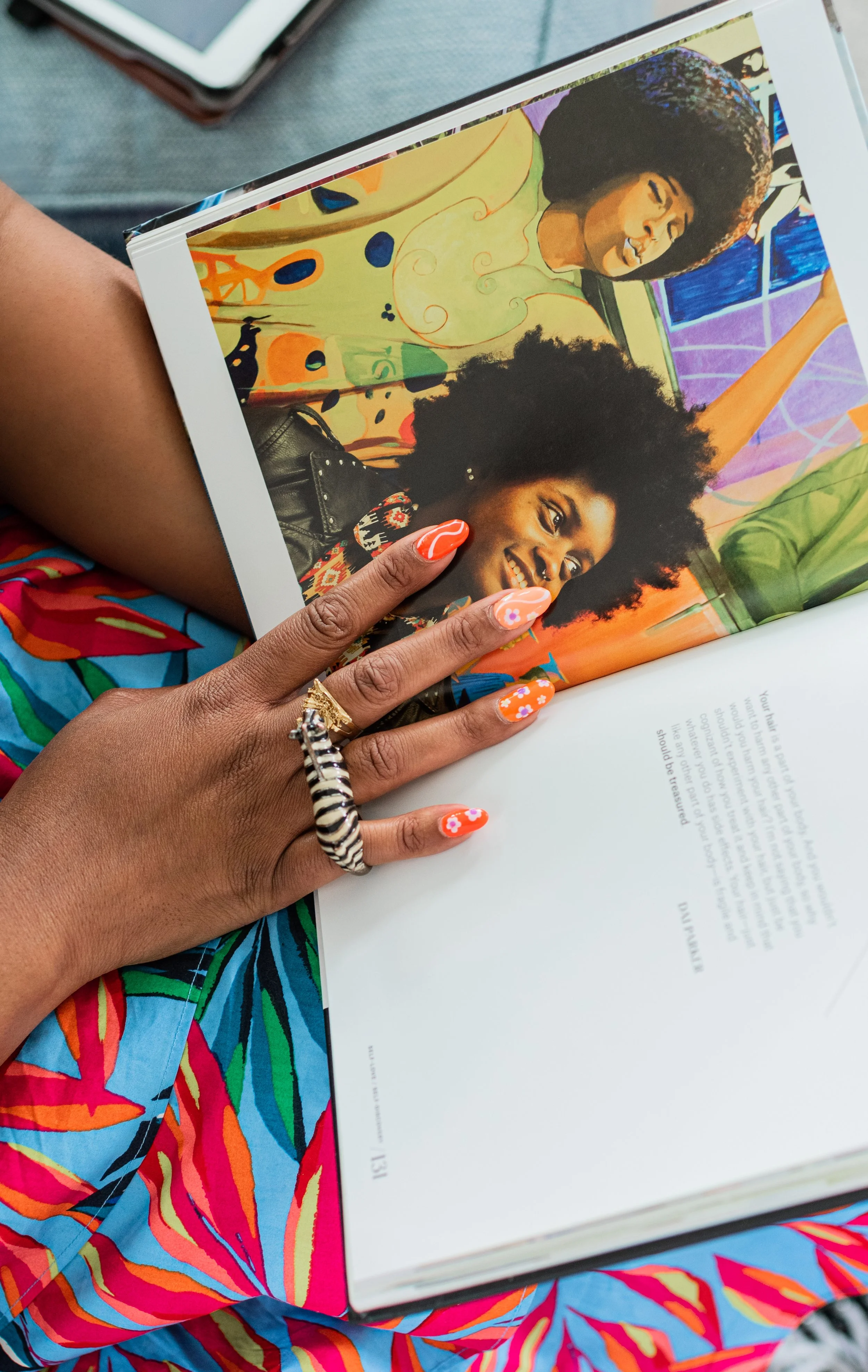 A person with decorated orange nails and rings, including a zebra-patterned ring, holds open a colorful magazine showing two women, one with curly hair and vibrant art in the background.