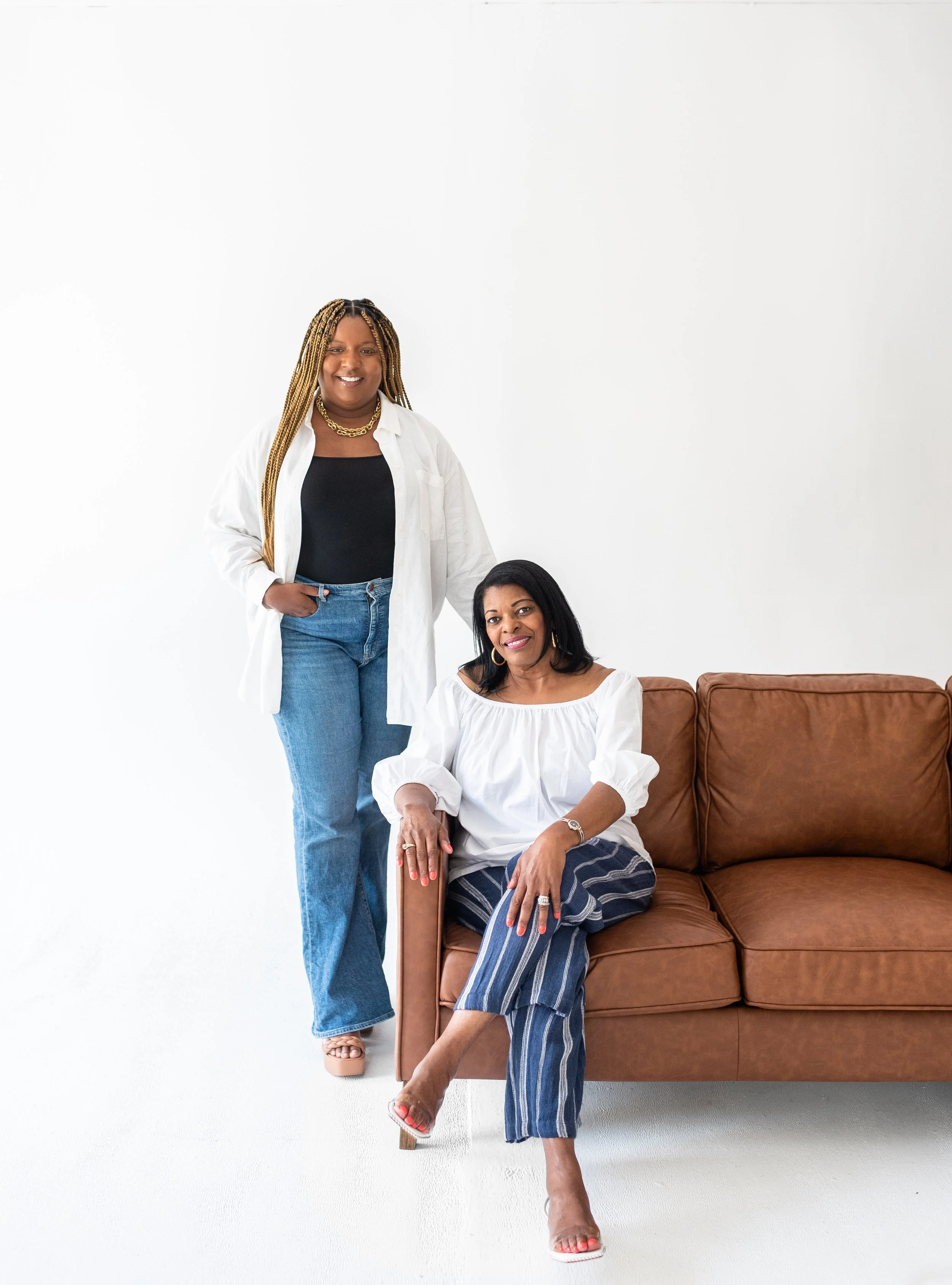 Two women posing in a bright, minimalistic room with a brown couch. One is standing with her hand in her pocket, and the other is sitting on the couch. Both women are smiling and wearing fashionable outfits.