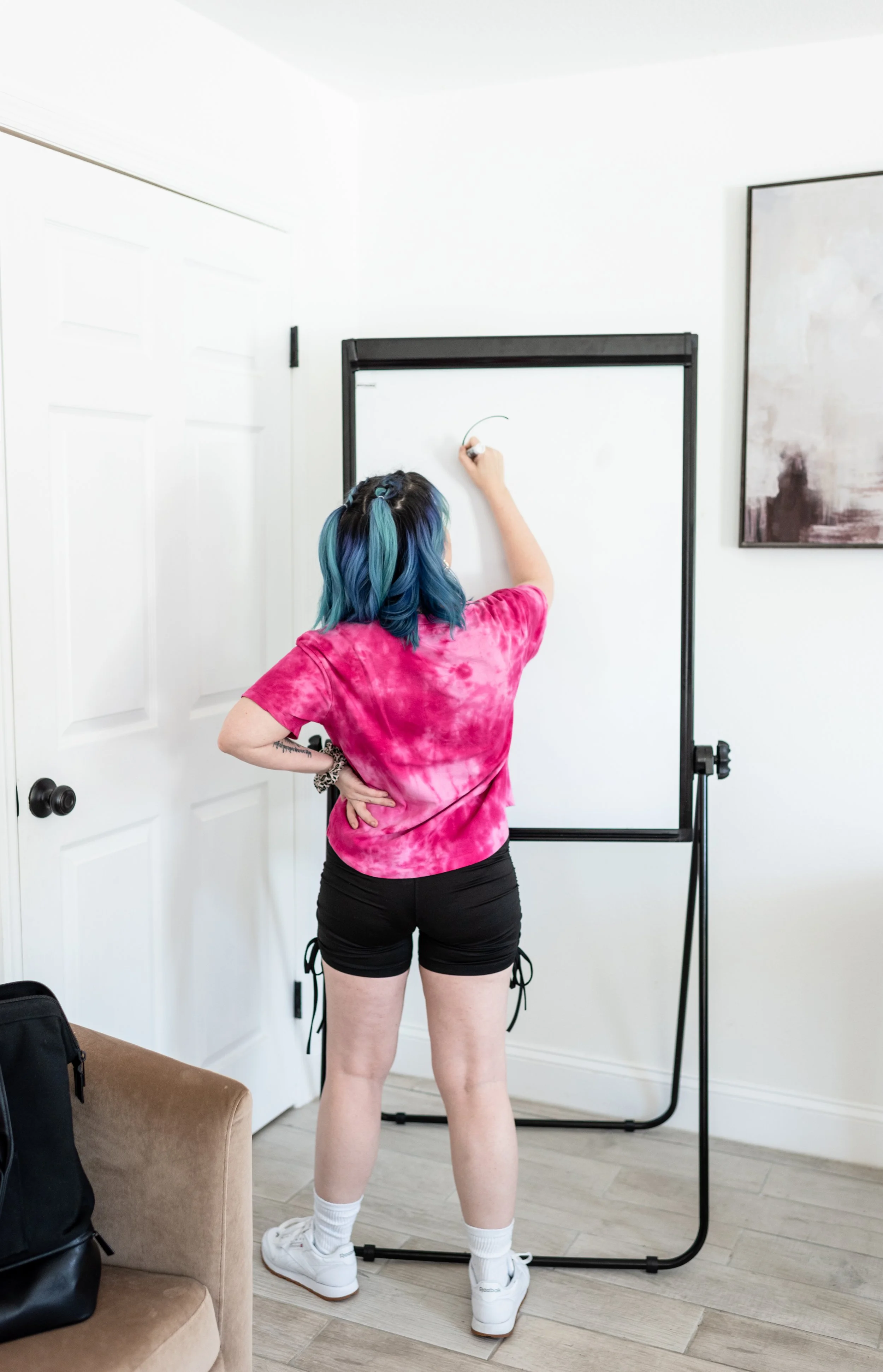 A person with blue and teal hair, wearing a pink tie-dye shirt and black shorts, drawing on a whiteboard in a room with white walls and a framed painting.