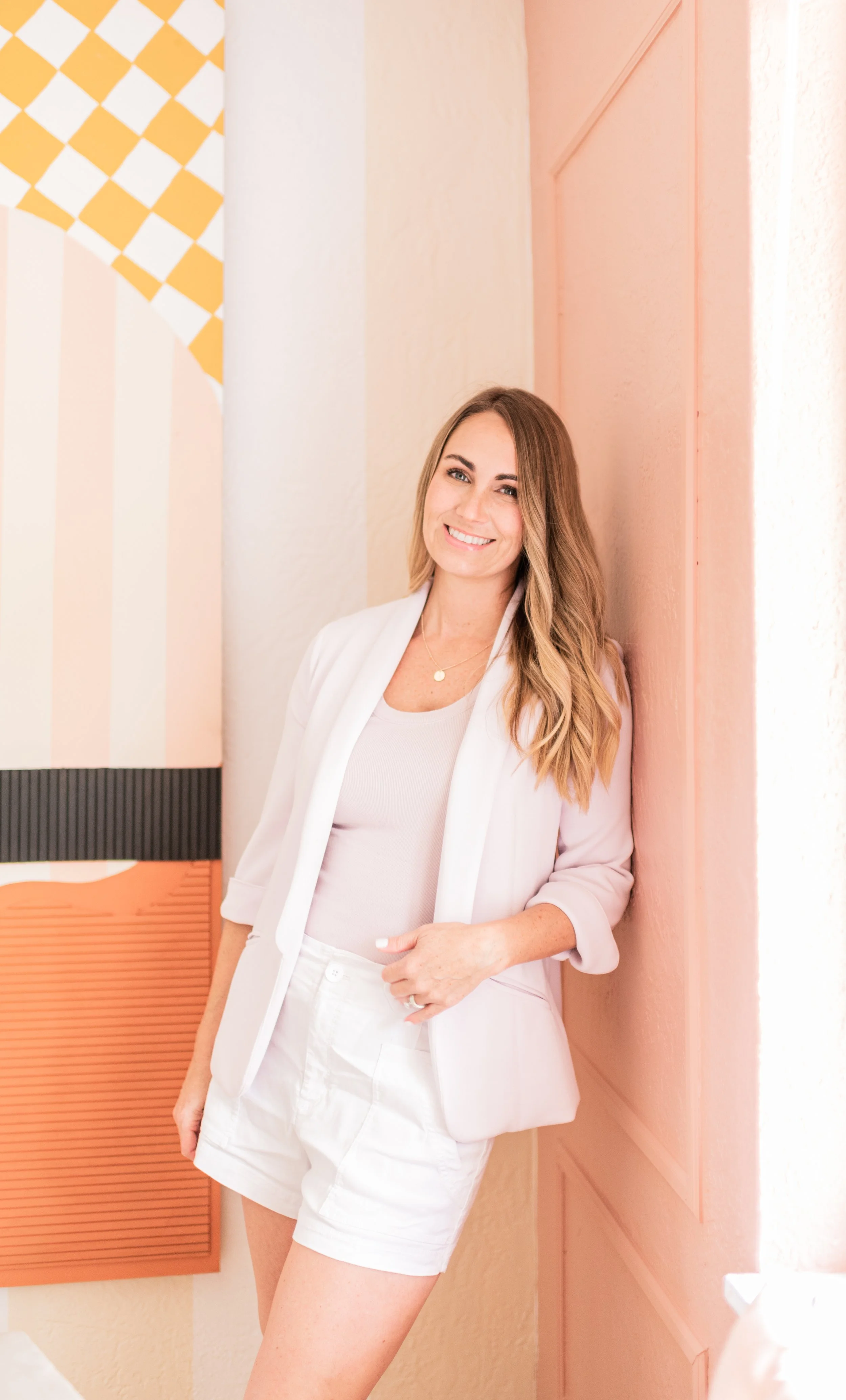 A woman with long blonde hair, smiling, dressed in a white blazer, matching top, and white shorts, leaning against a pink wall inside a brightly decorated room.