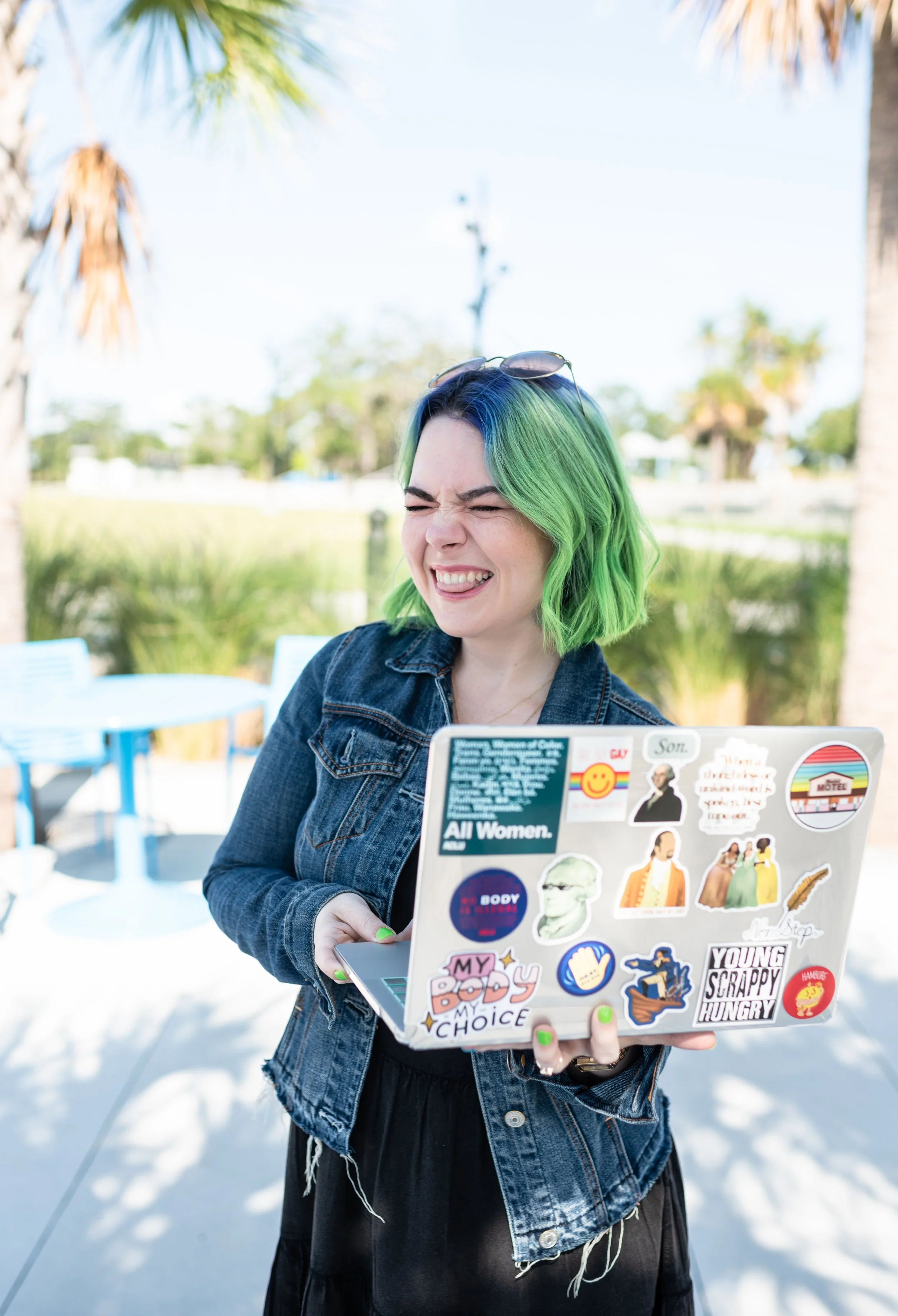 A woman with green and blue hair squinting and smiling while holding a laptop covered in colorful stickers, outdoors on a sunny day with palm trees and a lake in the background.
