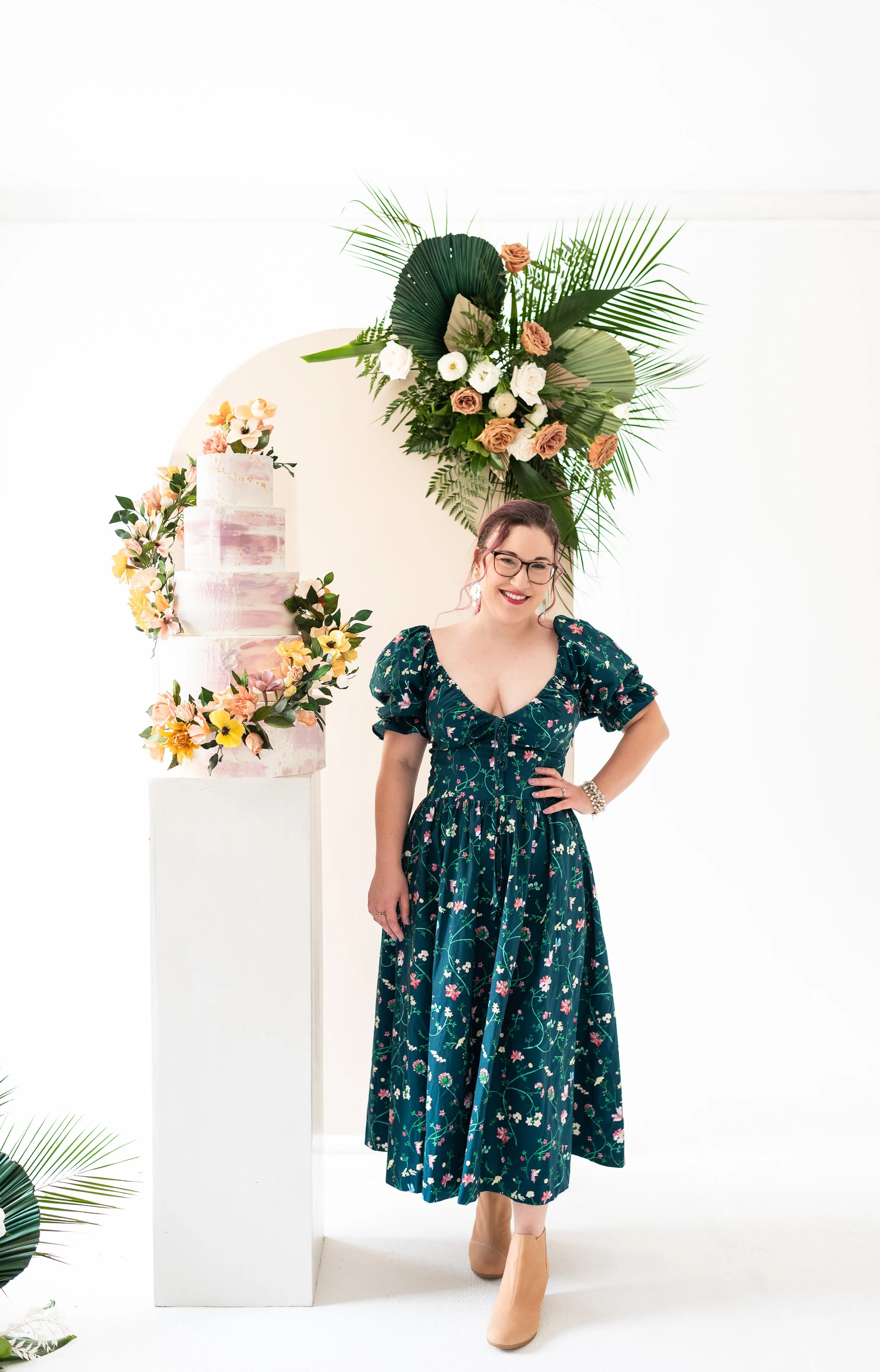 A woman in a floral dress standing next to a decorated cake and greenery arrangement at a celebration or event.