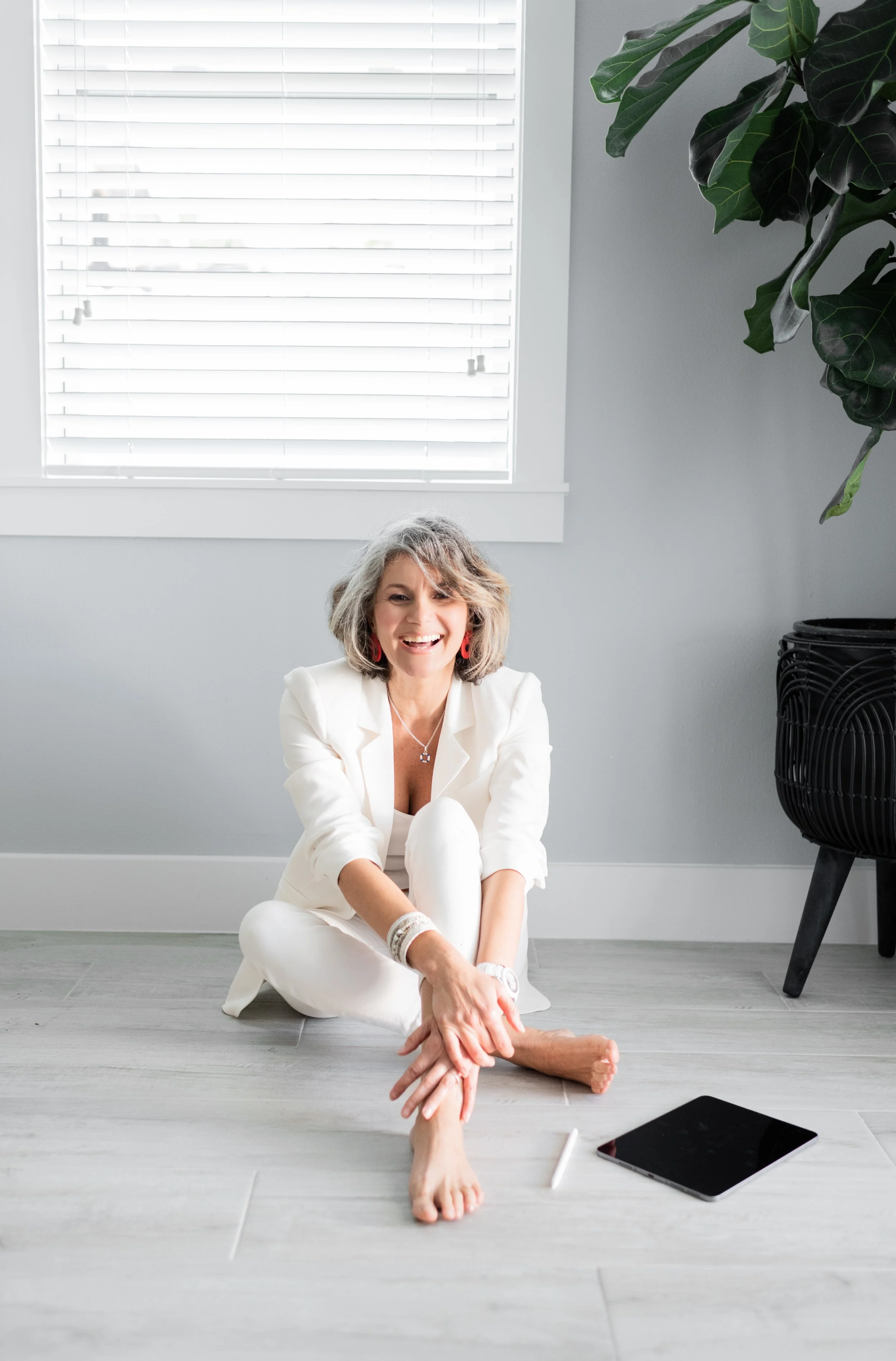 Smiling woman in white sitting on the floor with her legs crossed, near a window with closed blinds, a tablet and stylus on the floor, and a large potted plant in the background.