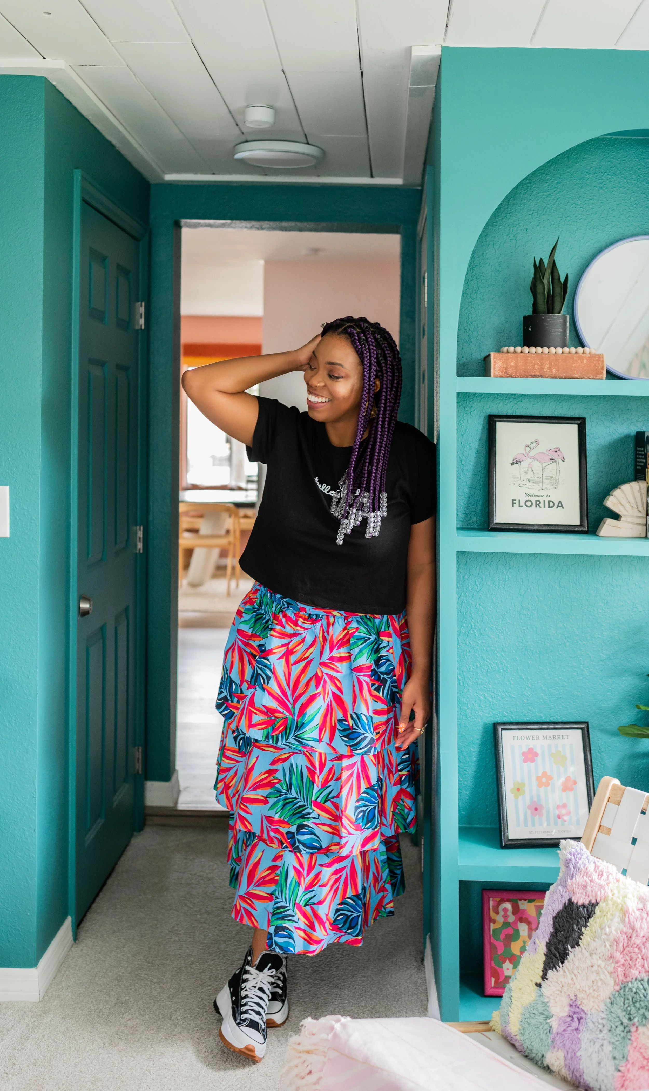 A woman with purple braided hair smiling and standing in a colorful, tropical-themed room.