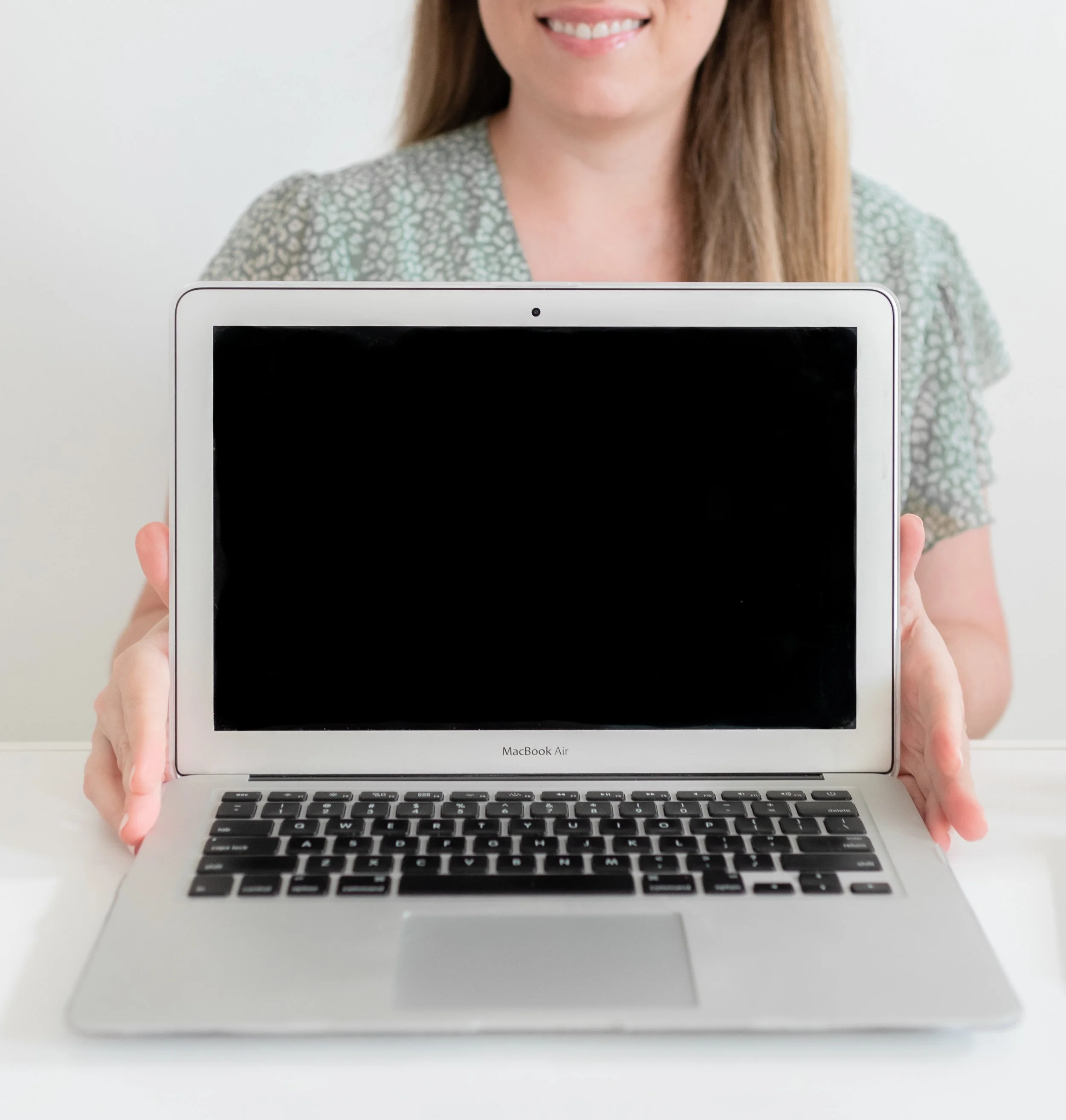 A woman in a patterned blouse holding a silver MacBook Air with a black screen, sitting at a white table.