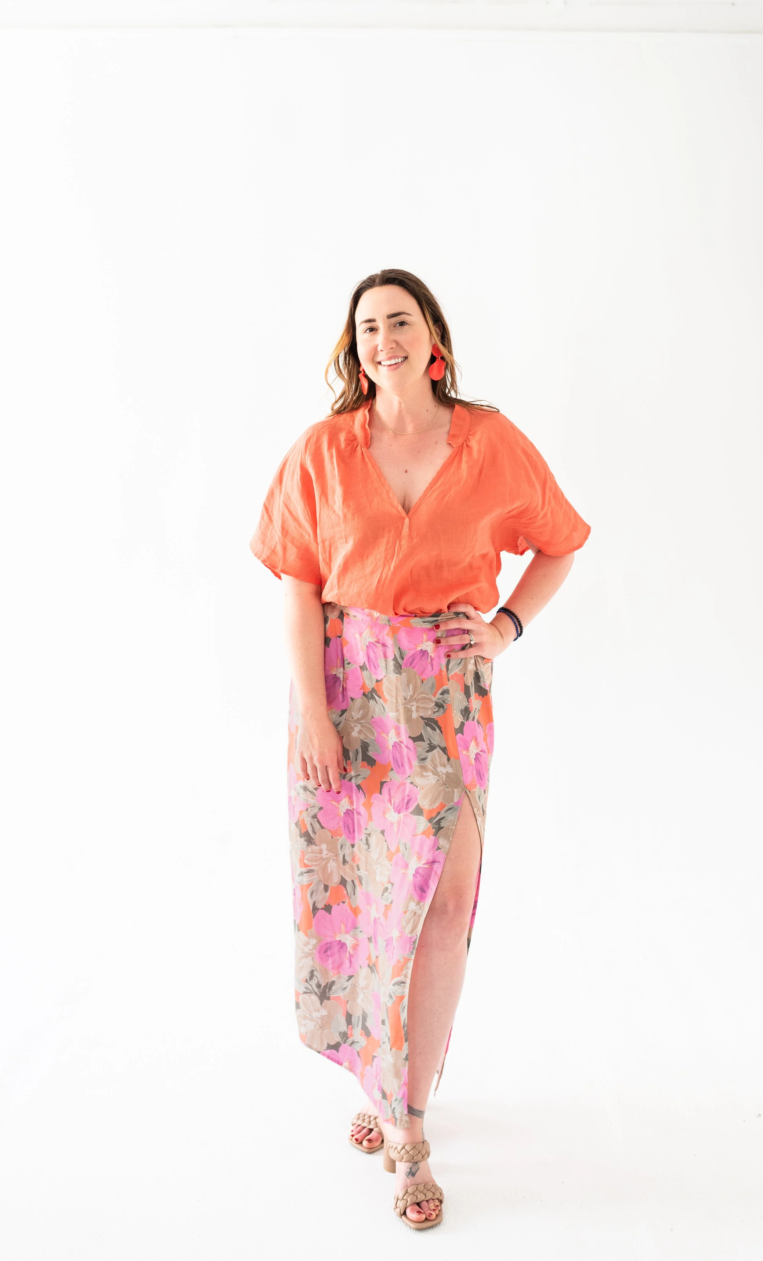 A woman wearing a bright orange top with a low V-neck and floral skirt with pink, purple, and beige flowers, standing against a plain white background, smiling with one hand on her hip and the other hanging by her side.