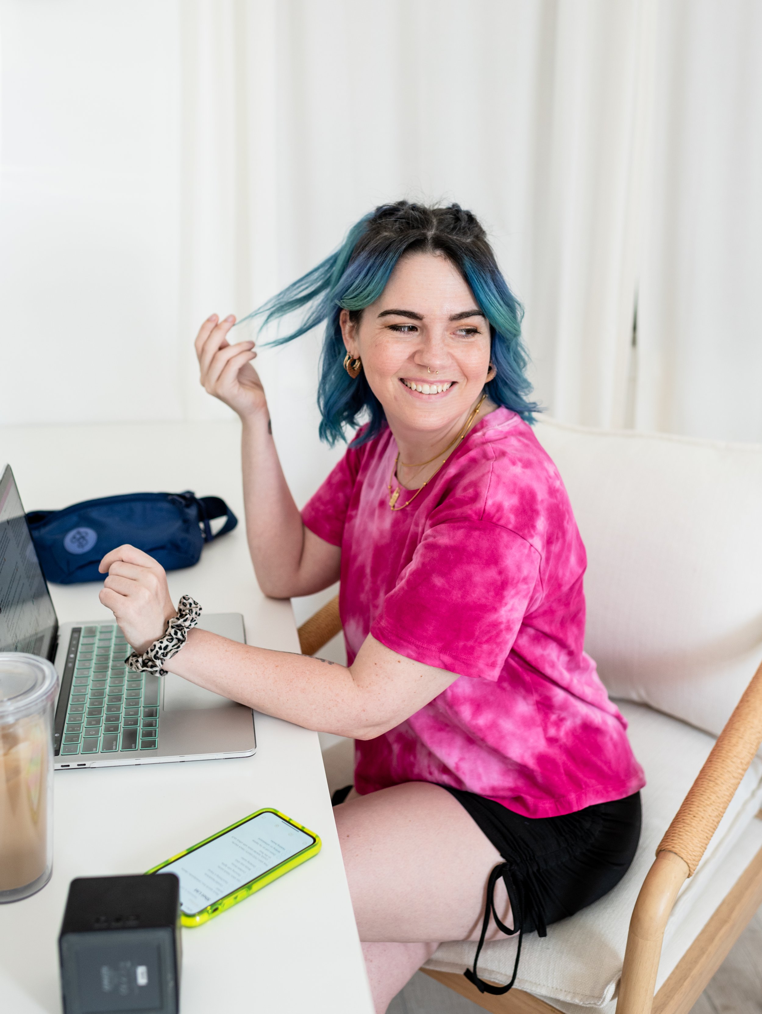A woman with blue and green hair smiling while sitting at a desk with a laptop, phone, and drinks, wearing a pink tie-dye shirt and black shorts.