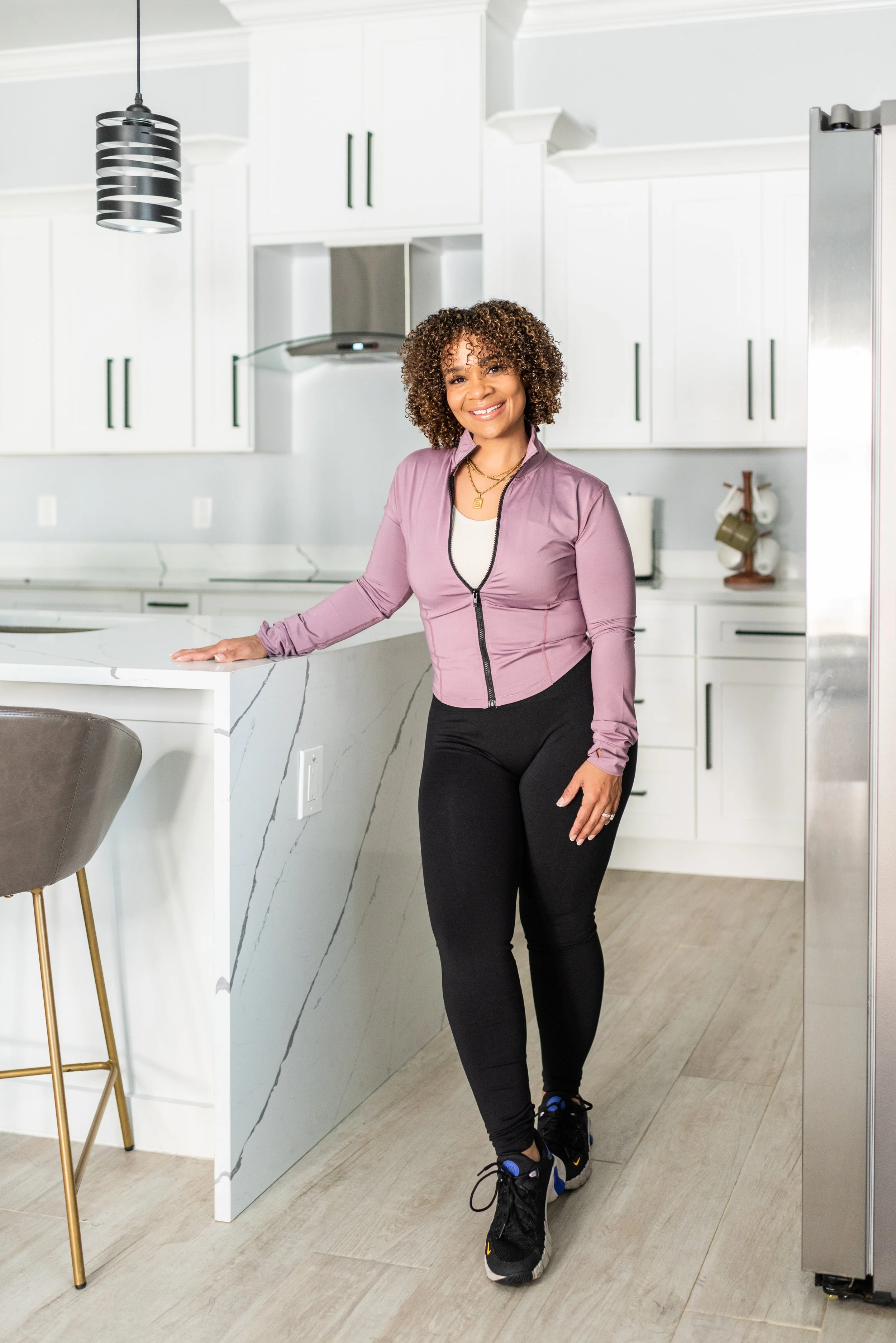 A woman smiling in a modern white kitchen, dressed in workout attire with black leggings, a light purple zip-up jacket, and sneakers.