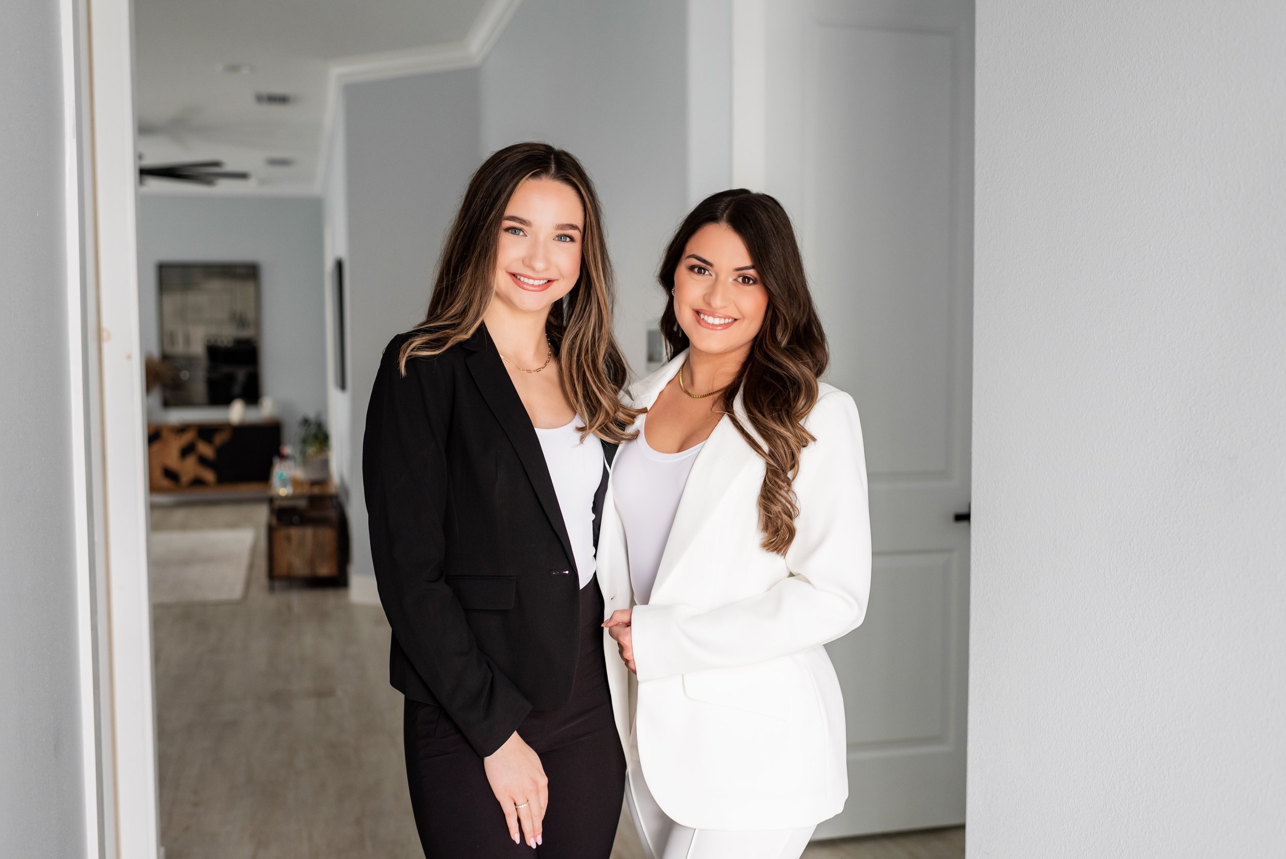 Two women dressed in business suits, smiling, standing together in a modern home interior.