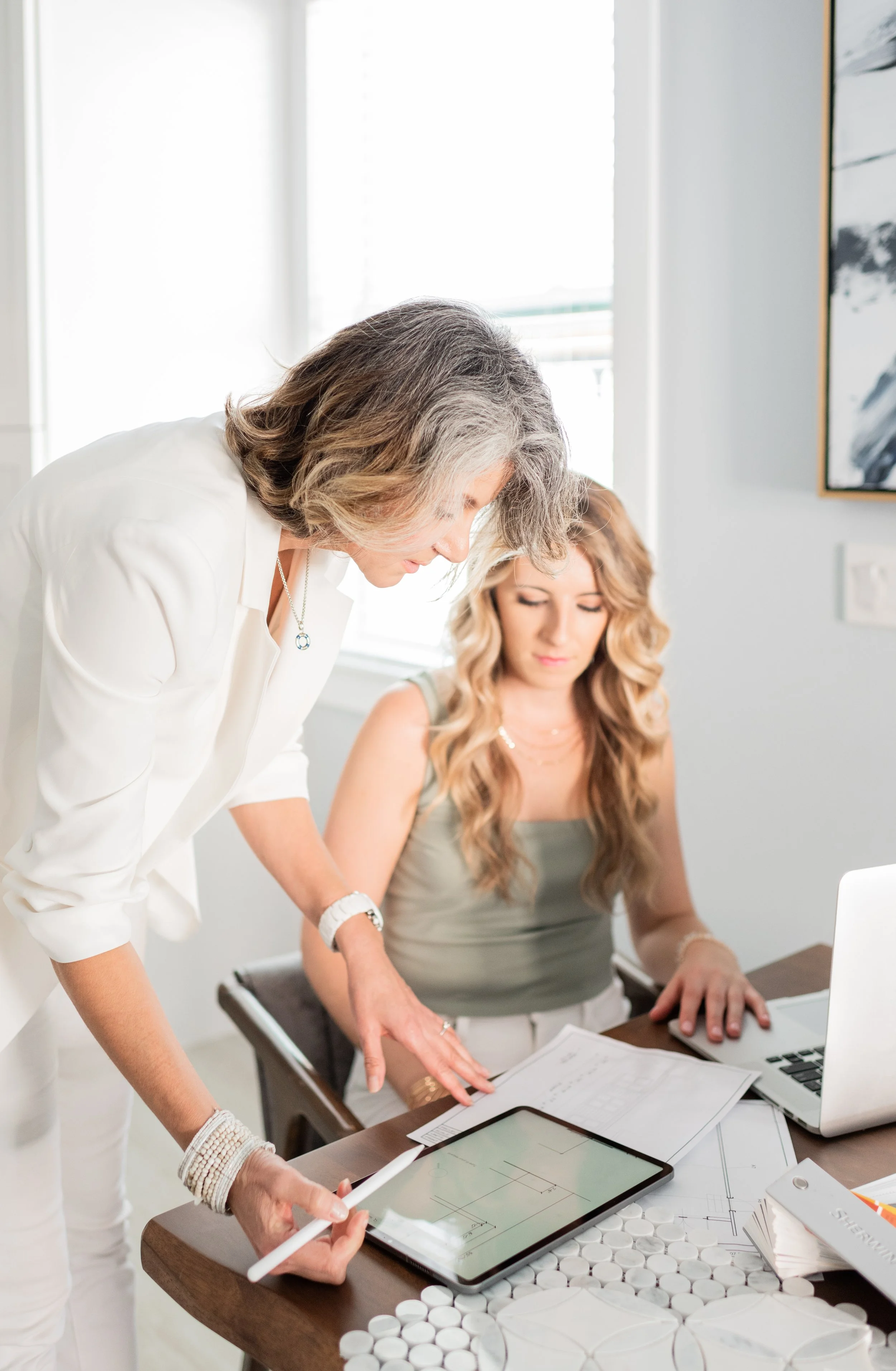 Two women are working together at a desk, looking at a tablet with a blueprint. The woman on the left is standing, in white, and the woman on the right is seated, with a laptop and papers.