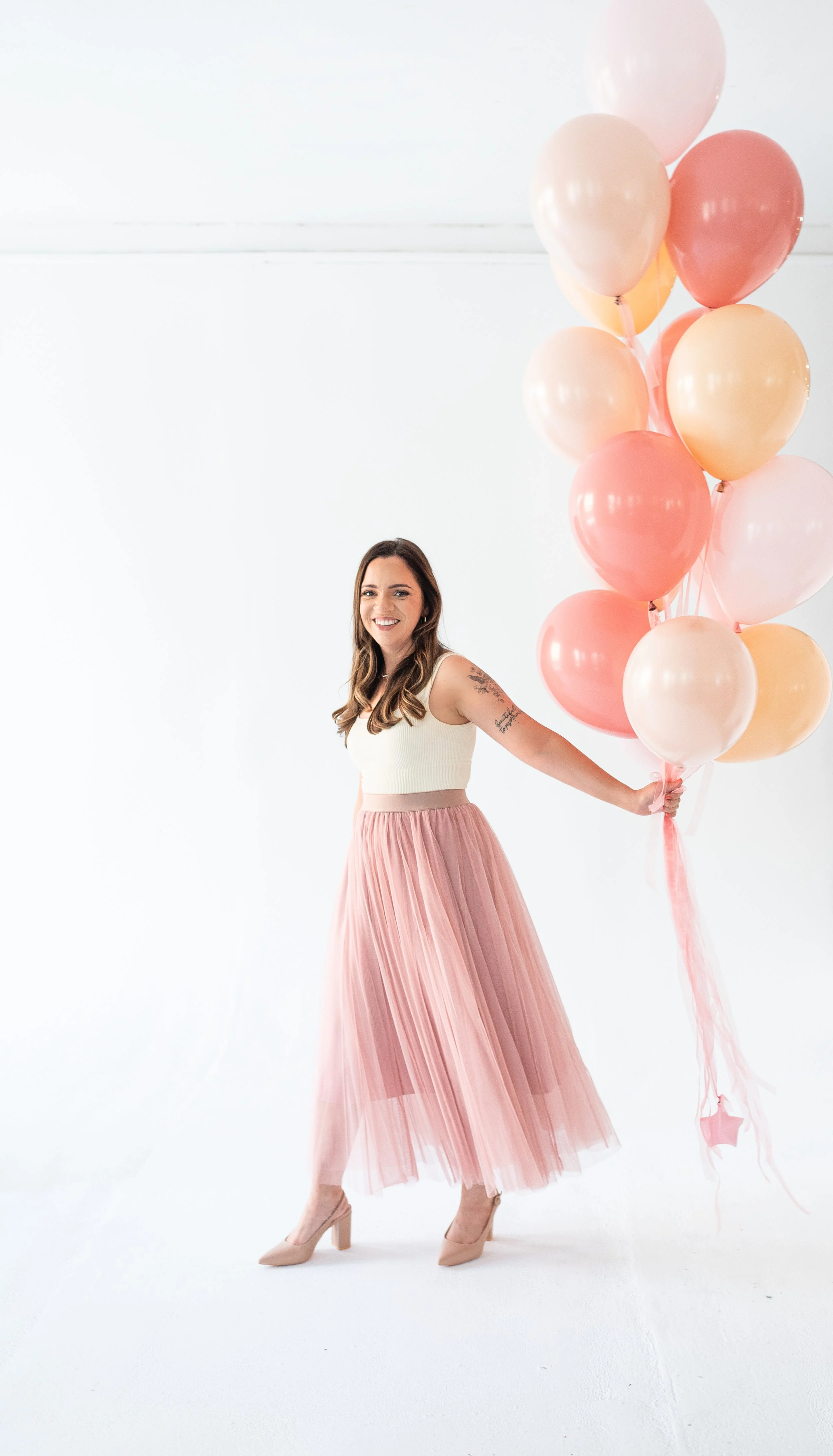 Woman in a white crop top and pink pleated skirt holding pink, peach, and white balloons in a white studio.