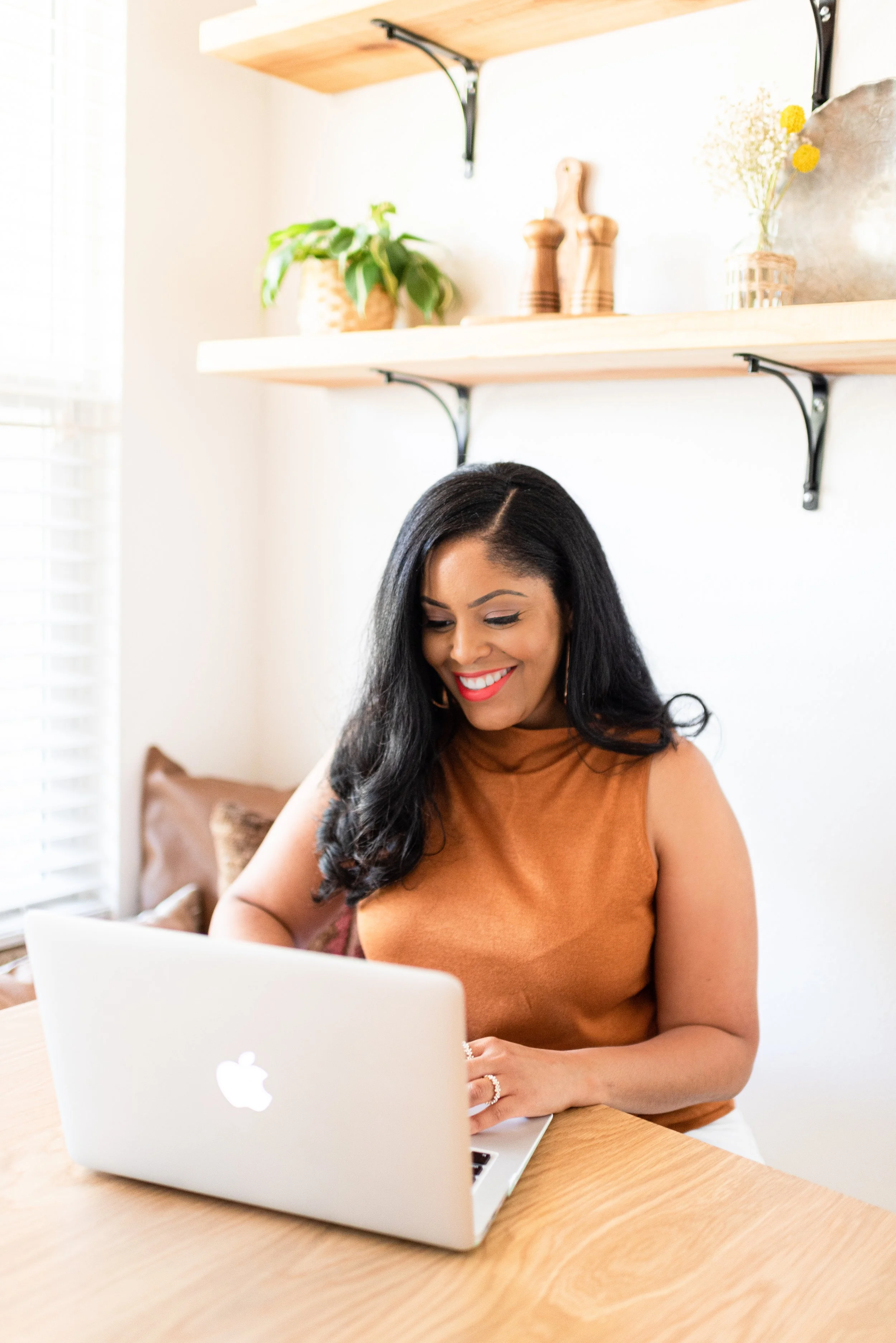 A woman with long black hair, wearing an orange sleeveless top, sitting at a wooden table and smiling while looking at her silver laptop, in a bright room with open shelves holding decorative items in the background.