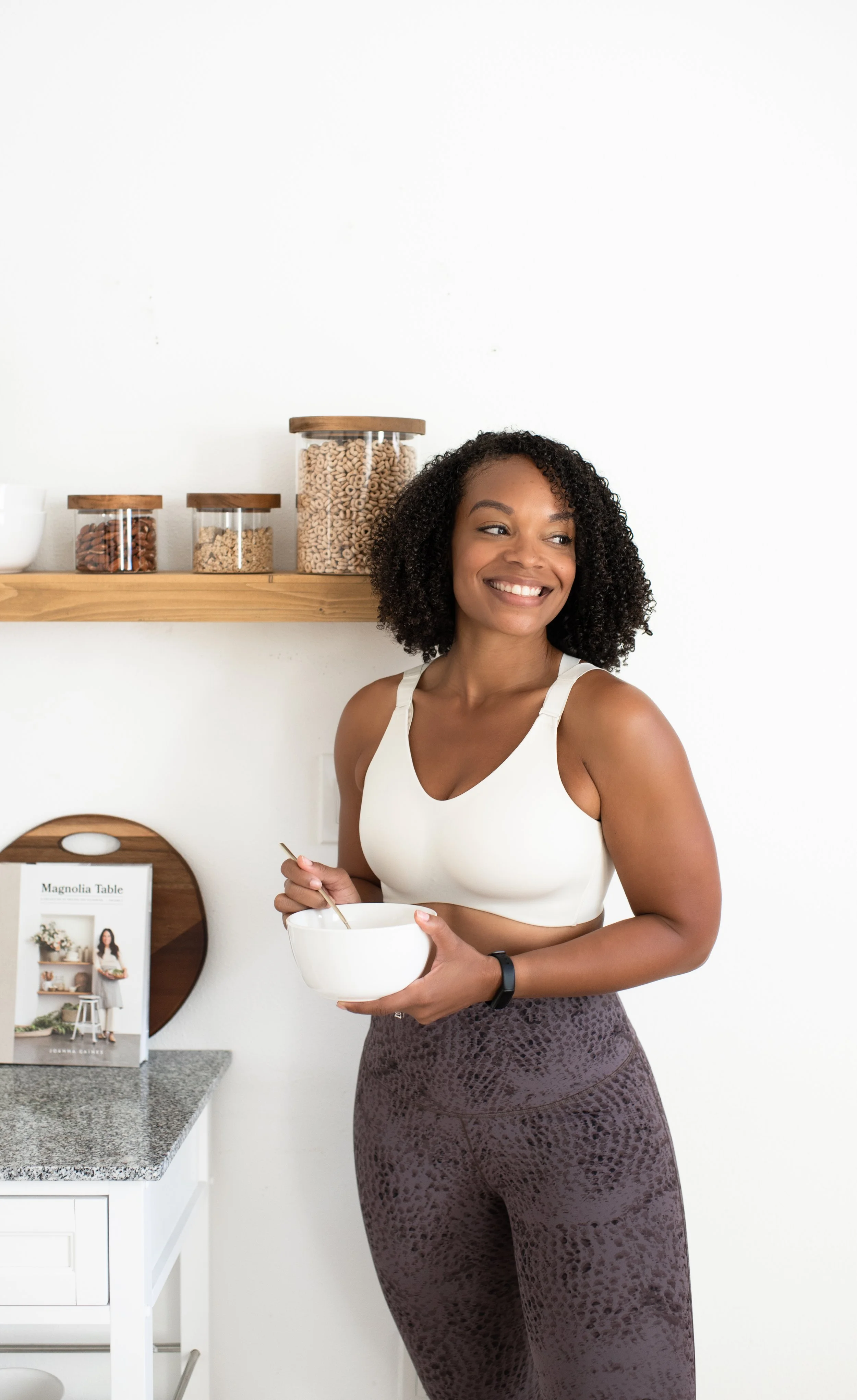A woman with curly hair smiling in a kitchen, holding a white bowl and spoon, wearing a white sports bra and patterned leggings.