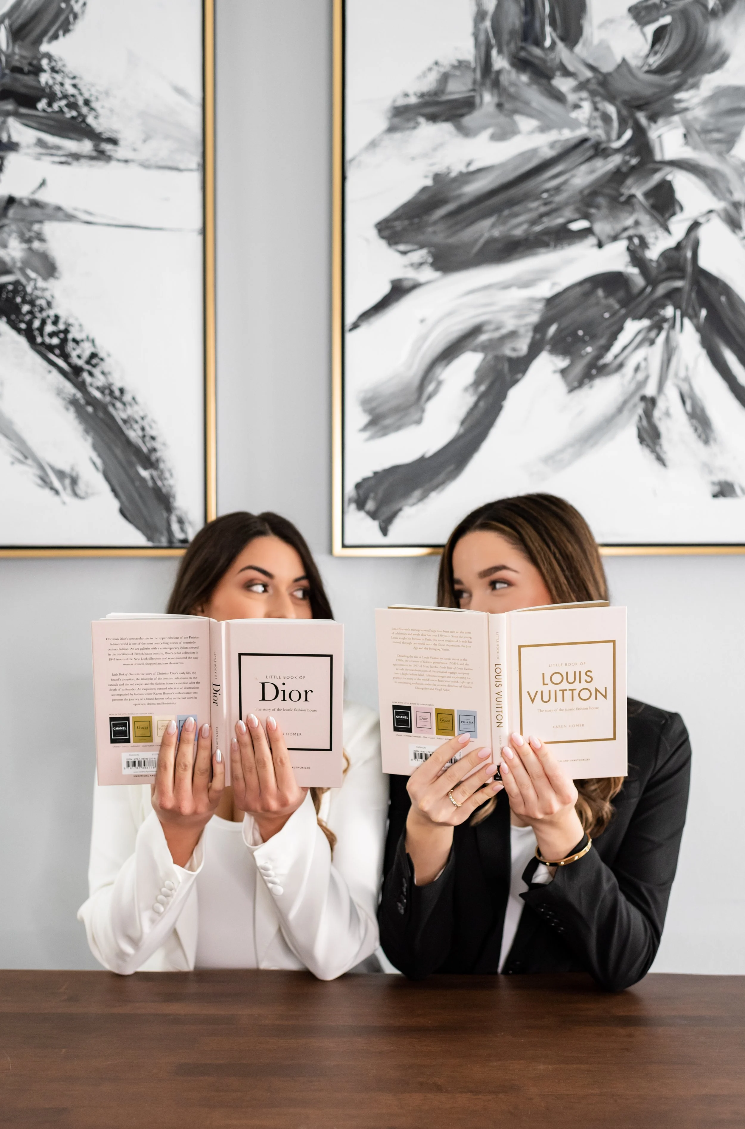 Two women sitting at a table holding fashion books and looking at each other, with abstract black and white artwork in the background.