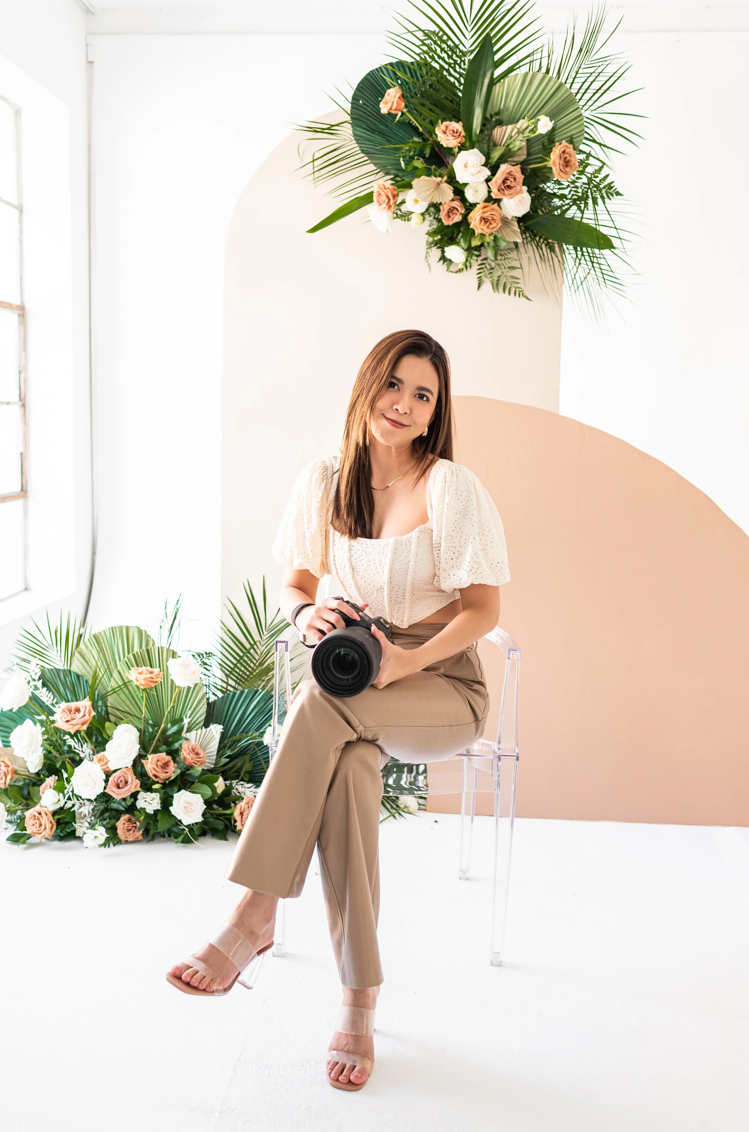 A woman sitting on a clear acrylic chair holding a camera, with floral arrangements and green foliage behind her, and a backdrop with a large floral and leaf arrangement.