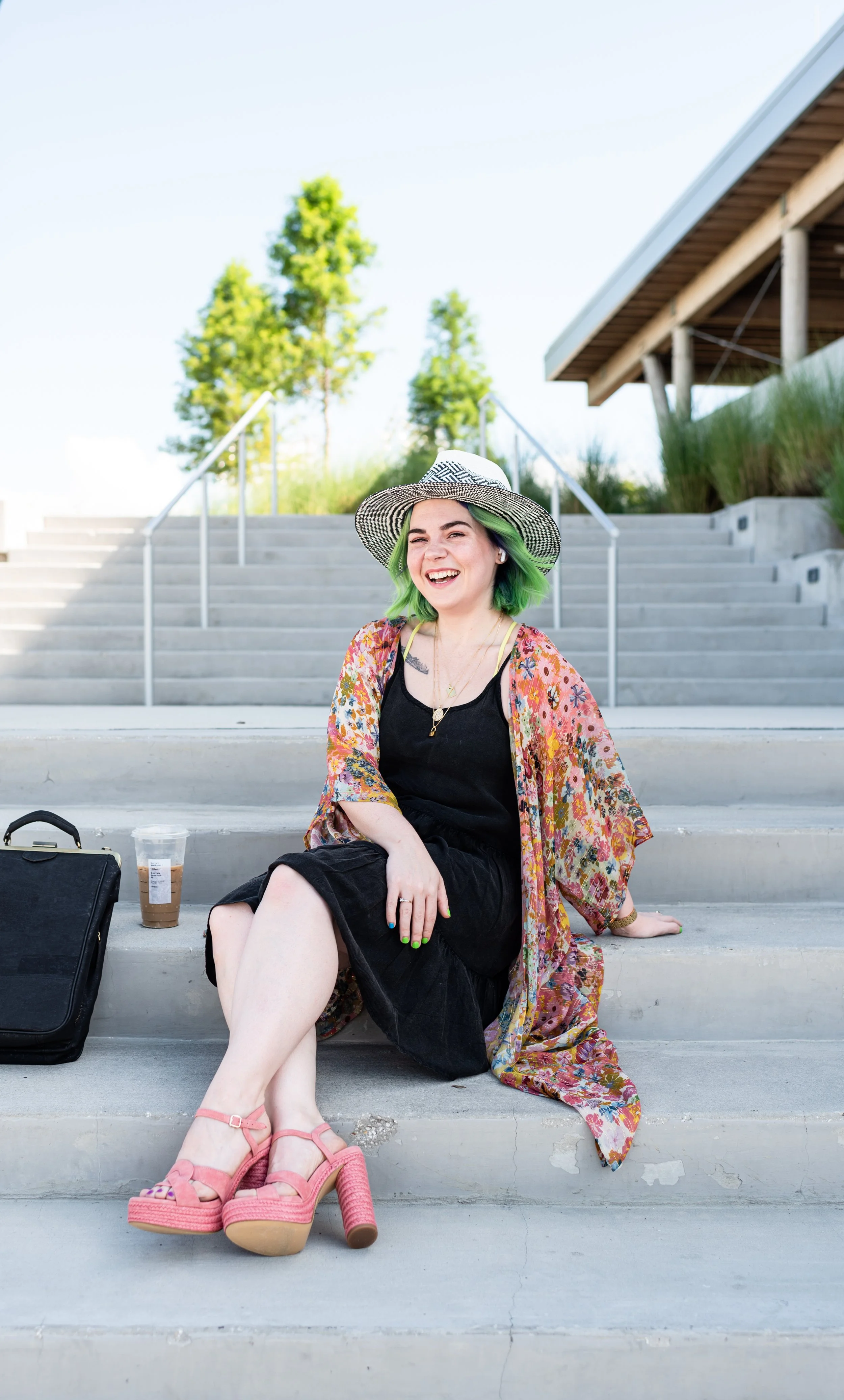 A young woman with green hair and a striped sun hat wearing a black dress and colorful shawl, sitting on concrete steps outdoors, smiling, with a coffee drink and black bag beside her.