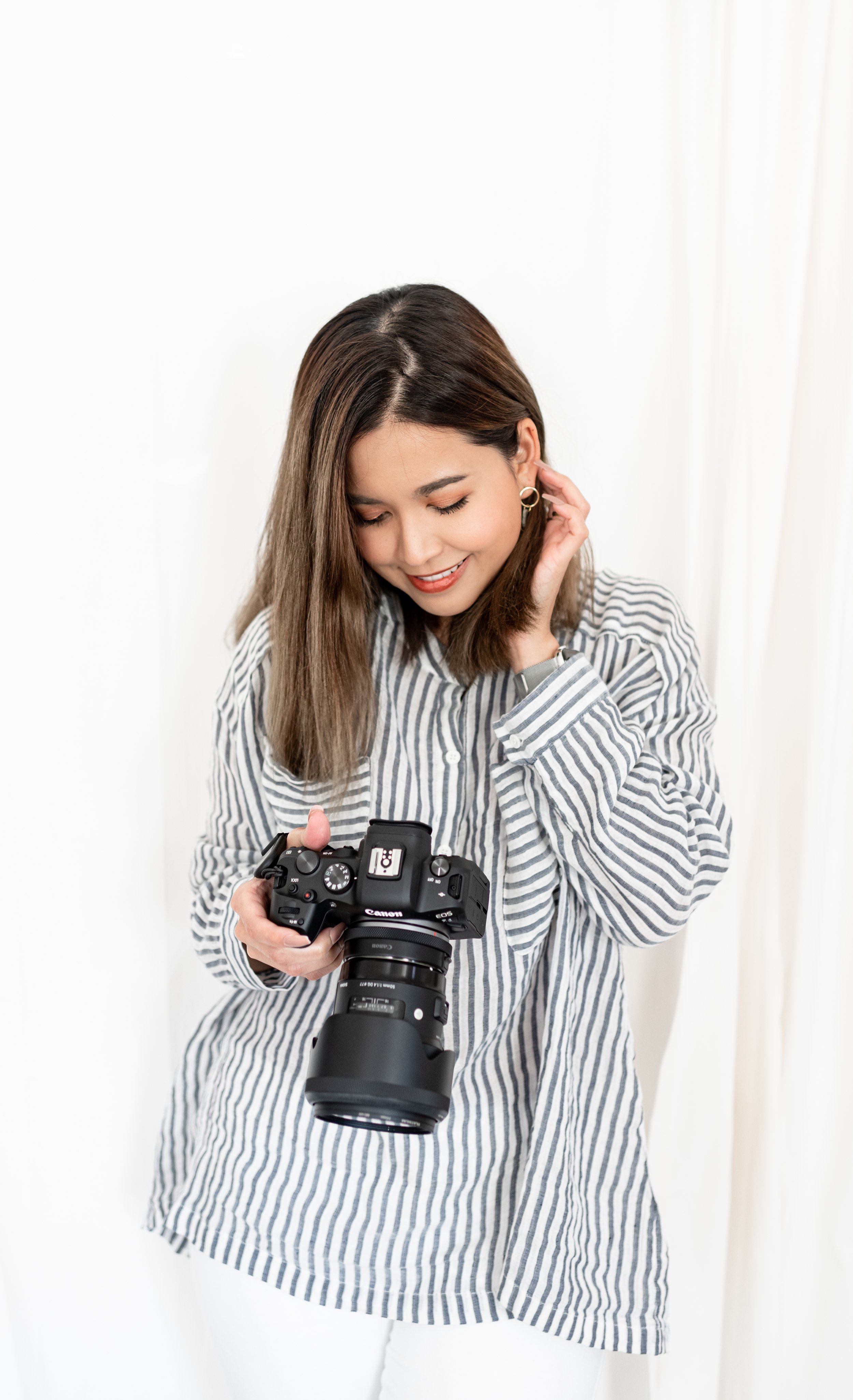 A woman with shoulder-length brown hair, wearing a striped shirt, smiling and looking down at her camera, holding a Canon camera with a large lens.