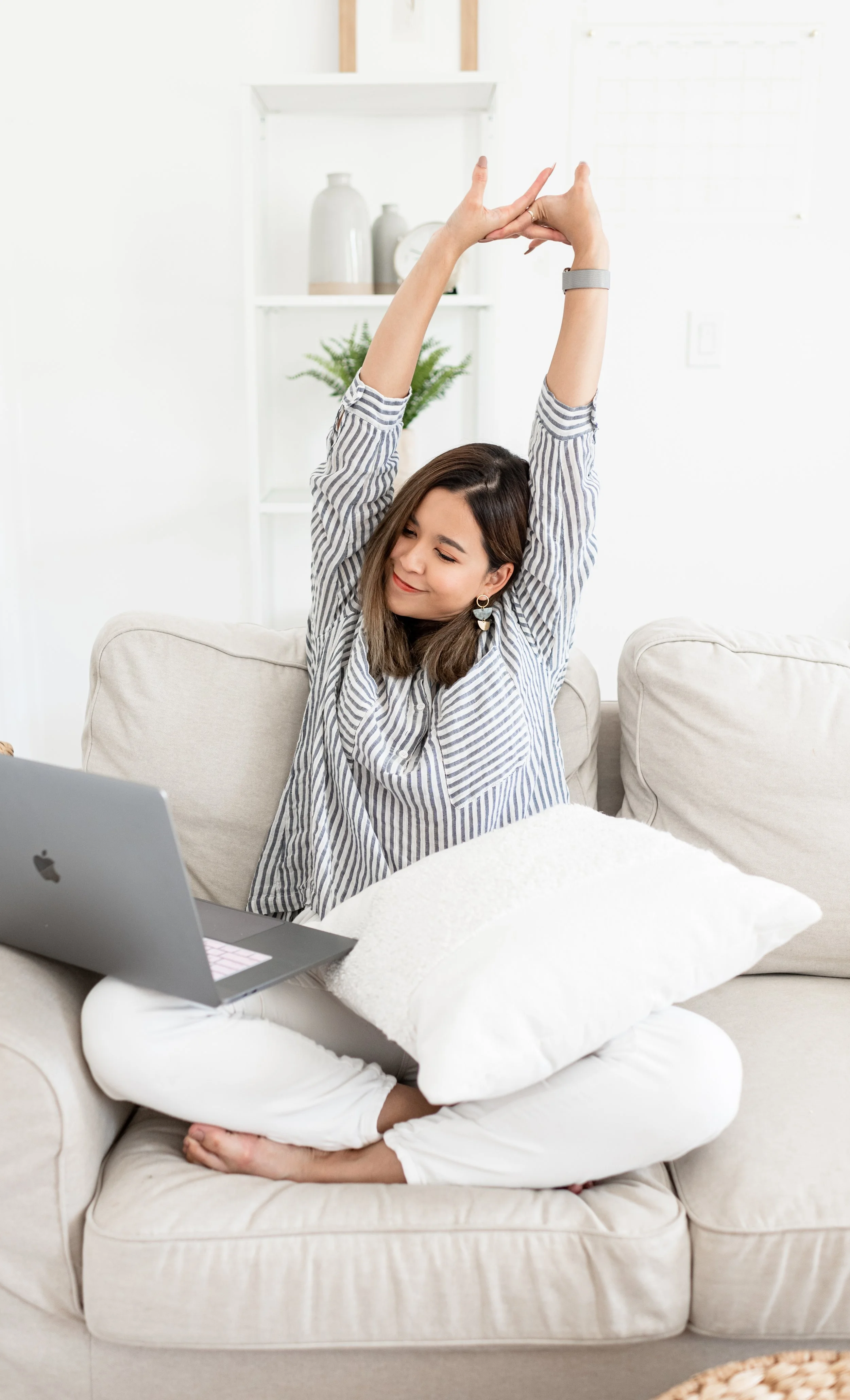 A woman sitting cross-legged on a beige sofa, stretching with her arms raised, in front of an open laptop, with a white cushion on her lap, in a bright living room.