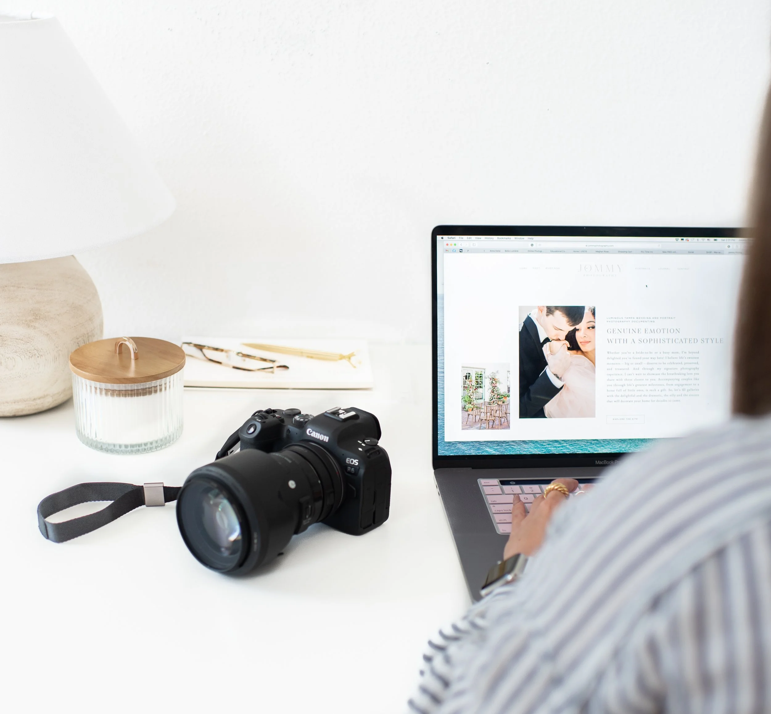 Person working on a MacBook laptop at a white desk with a Canon camera, a glass jar with a lid, a notepad, and eyeglasses nearby.