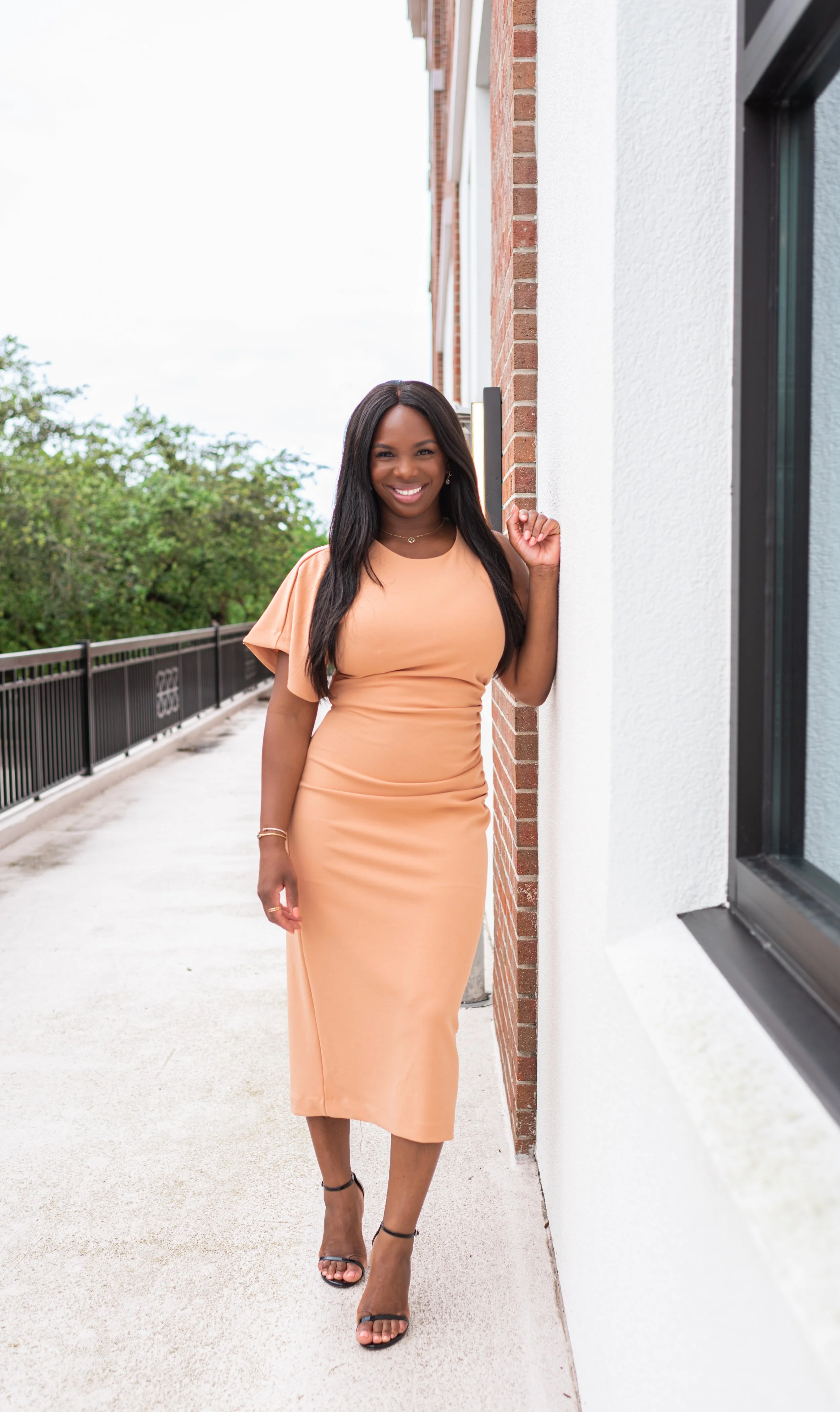 A woman in a peach dress and black high heels standing outside near a brick building, smiling at the camera.