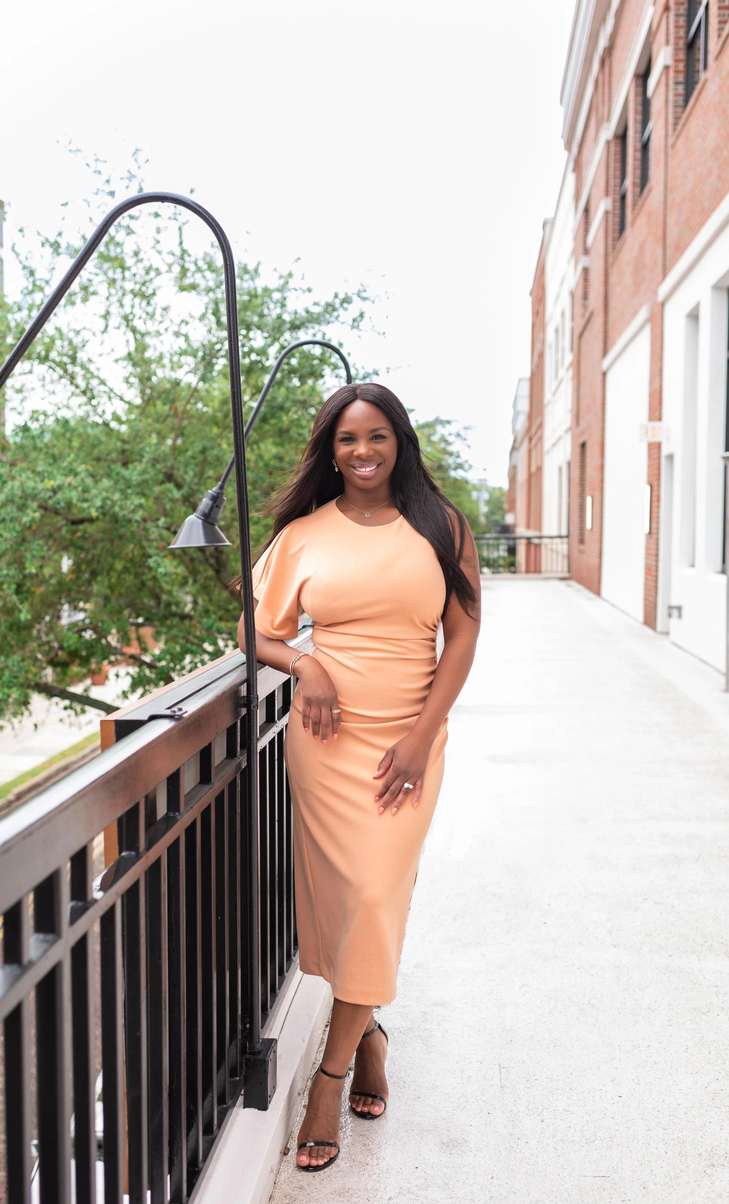 A woman in a peach-colored dress standing on an outdoor balcony railing, smiling.