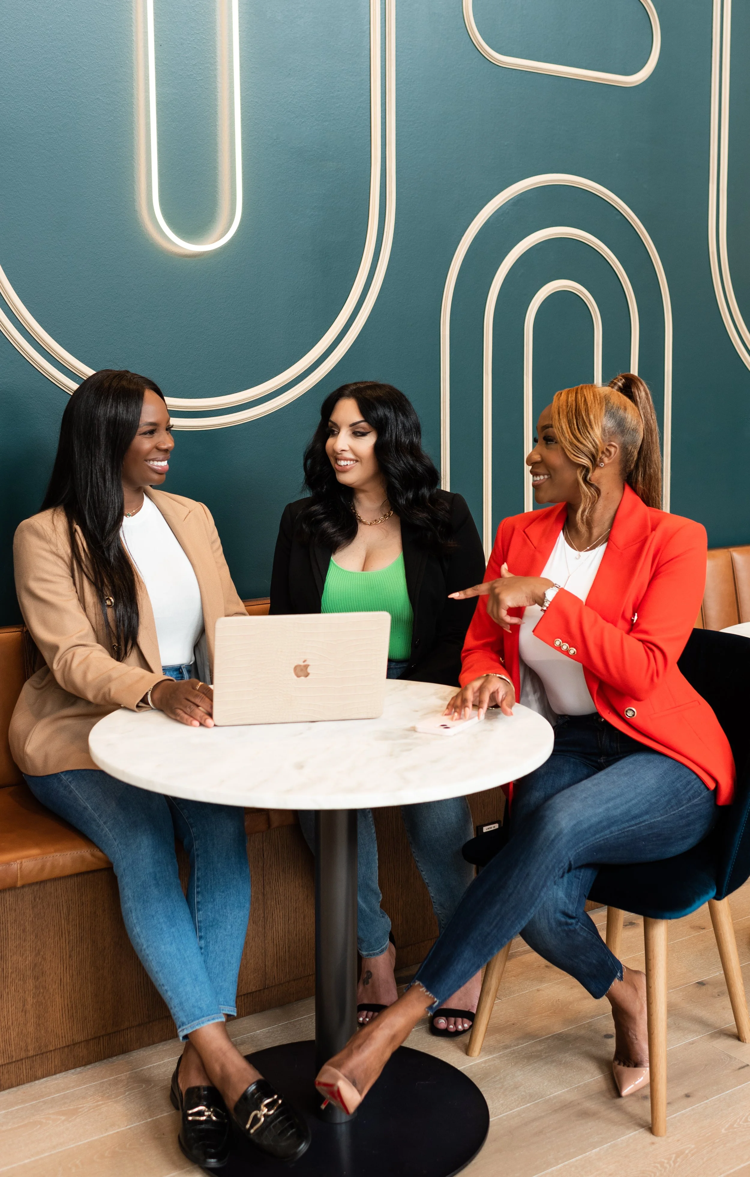 Three women sitting around a table in a coffee shop, engaging in conversation with one woman pointing and smiling.