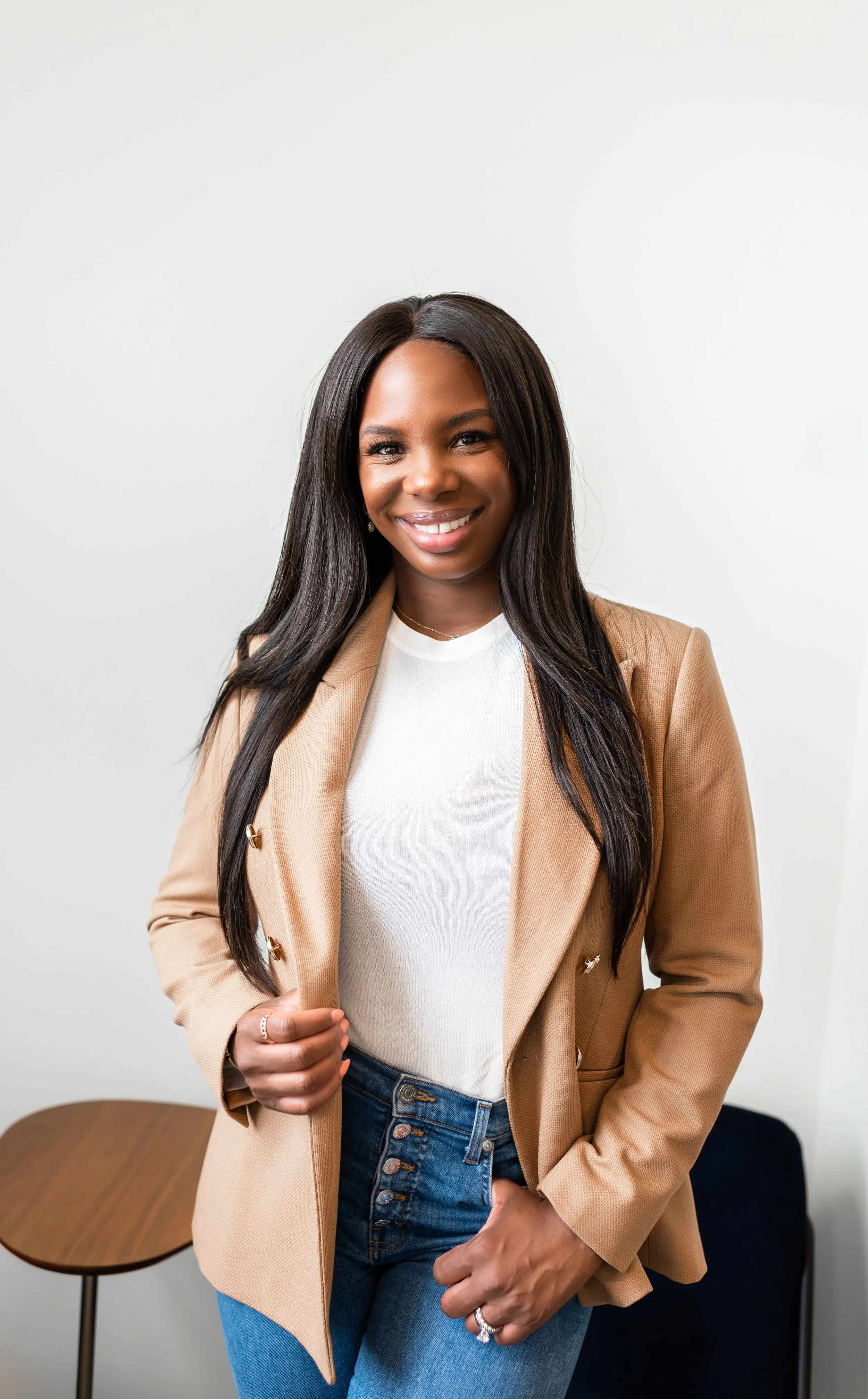 A woman with long black hair smiling, wearing a beige blazer over a white shirt, and blue jeans, standing in a room with white walls.