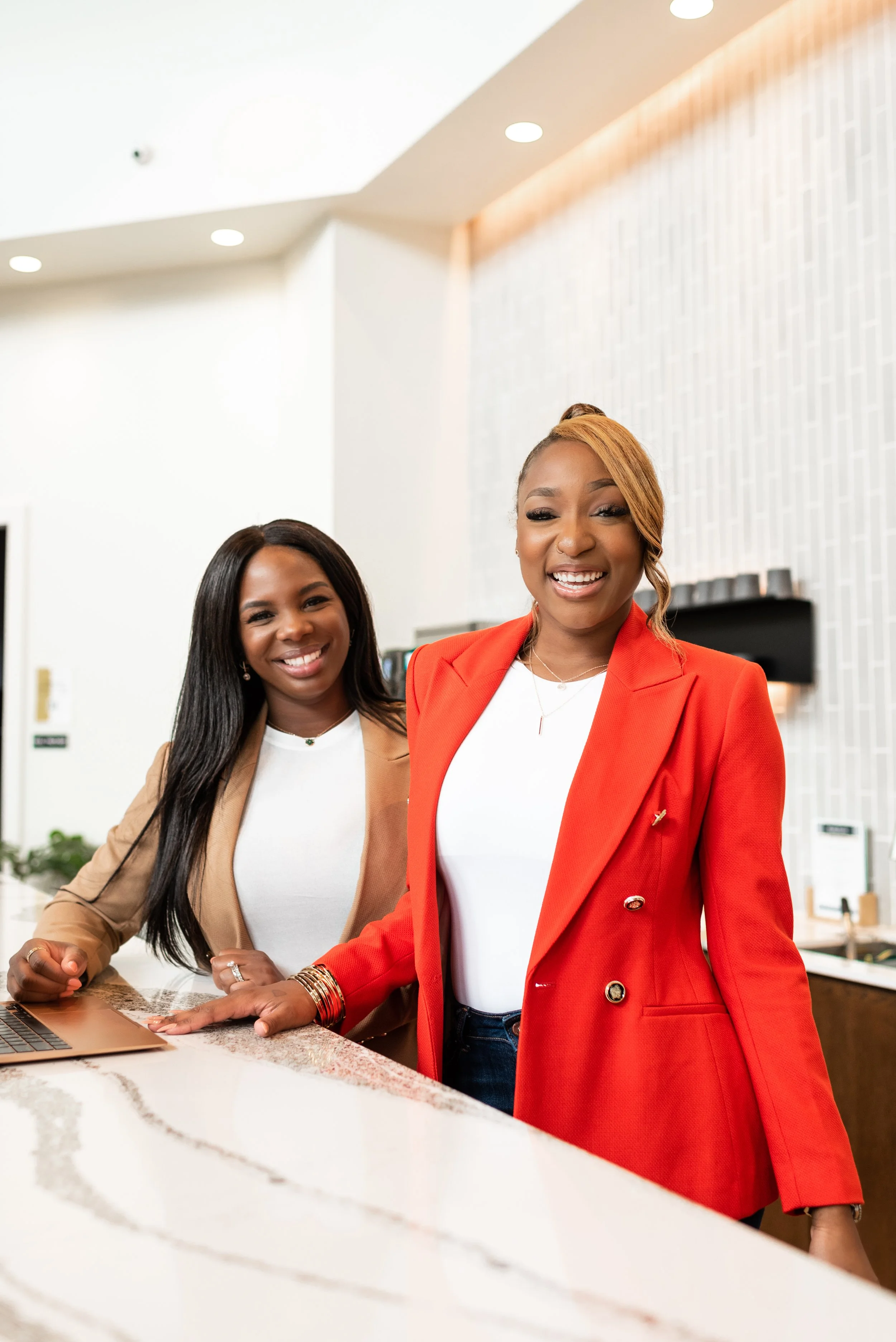 Two smiling women standing at a modern reception counter in an office lobby, one in a red blazer and the other in a beige blazer, with a laptop open on the counter.