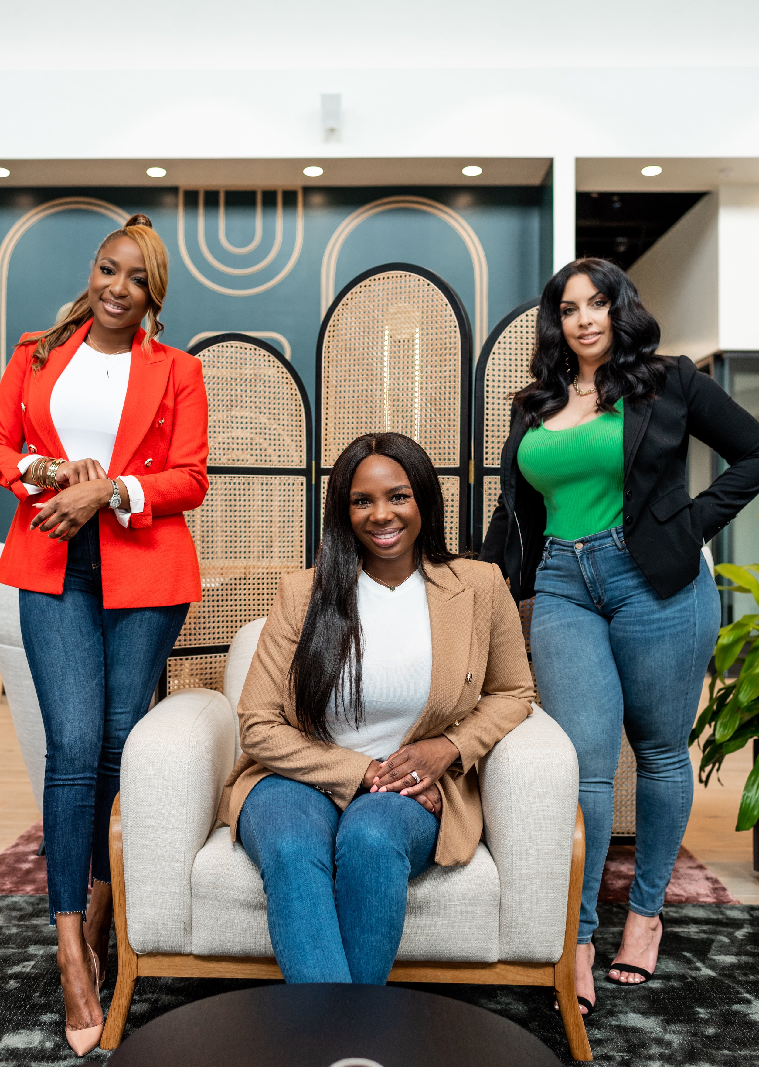 Three women posing in a modern office space. One woman is sitting on a beige armchair, and two women are standing behind her. The woman on the left is wearing a red blazer and jeans, and the woman on the right is wearing a black blazer with a green t
