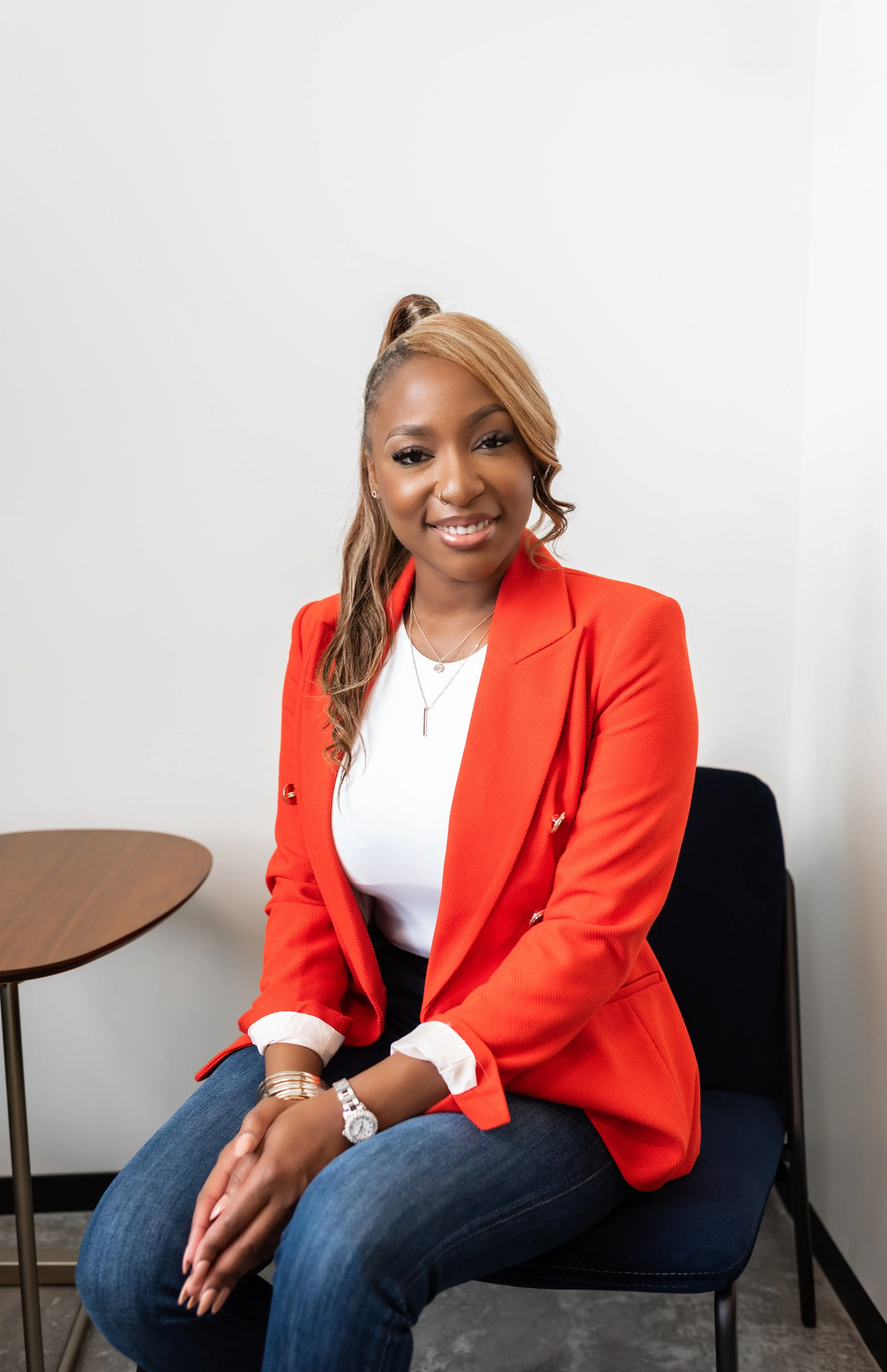 A woman with tan hair styled in a side ponytail, wearing a red blazer, white top, and blue jeans, sitting on a black chair in a room with a white wall and a small wooden table.