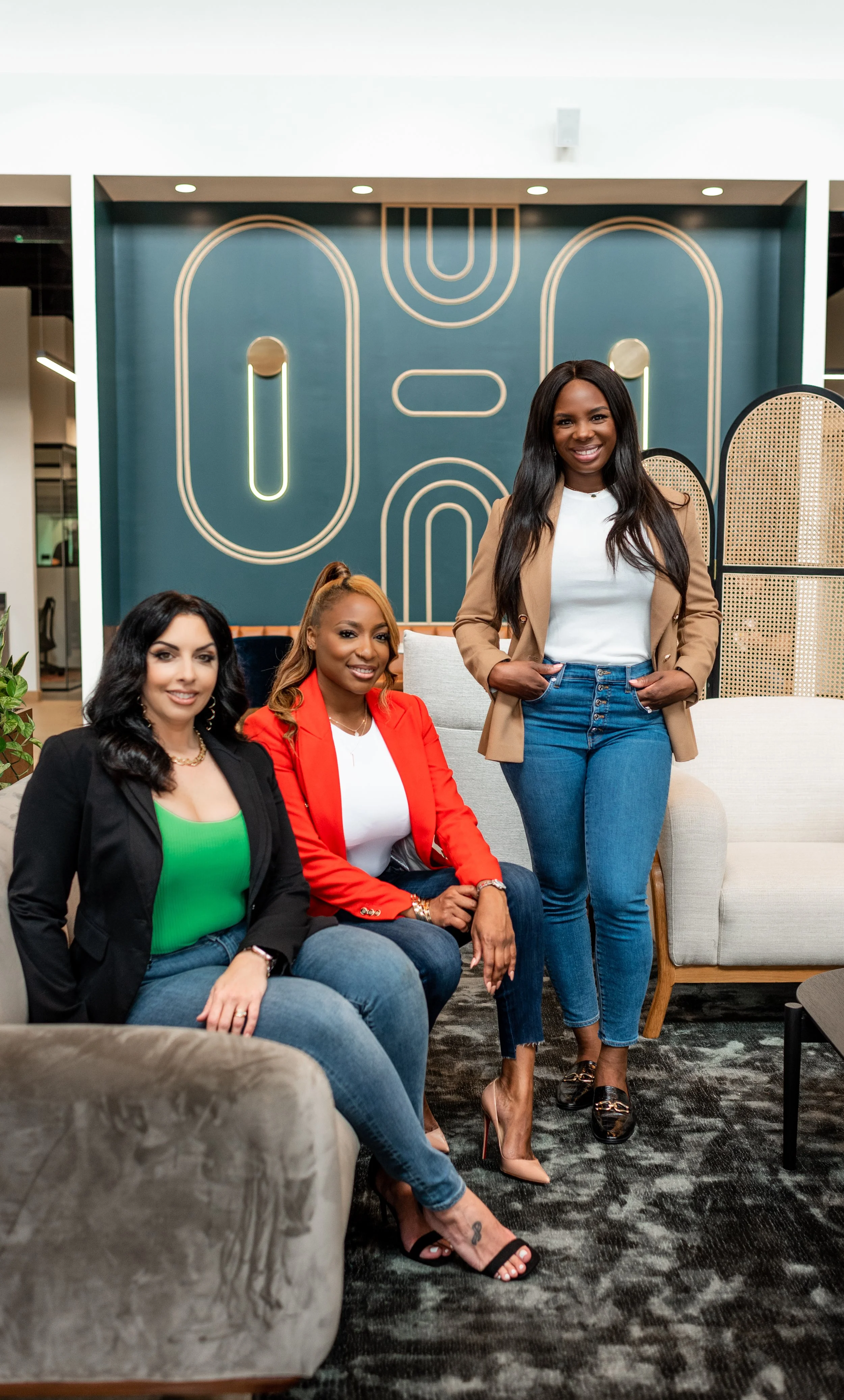 Three women sitting and standing on sofas in a modern office lobby with geometric wall art.